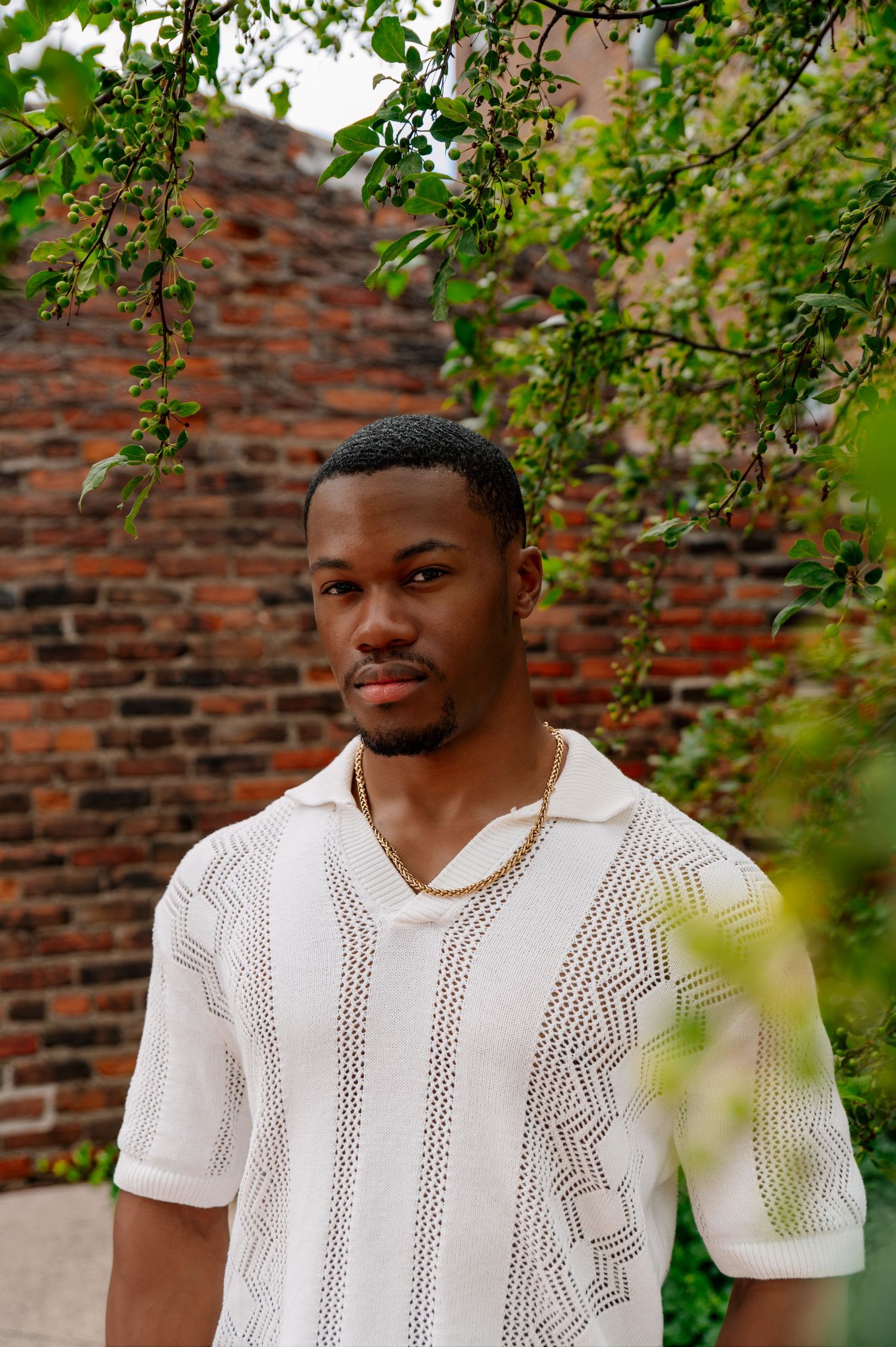 Portrait of man in white knit shirt against rustic brick wall with overhanging foliage.