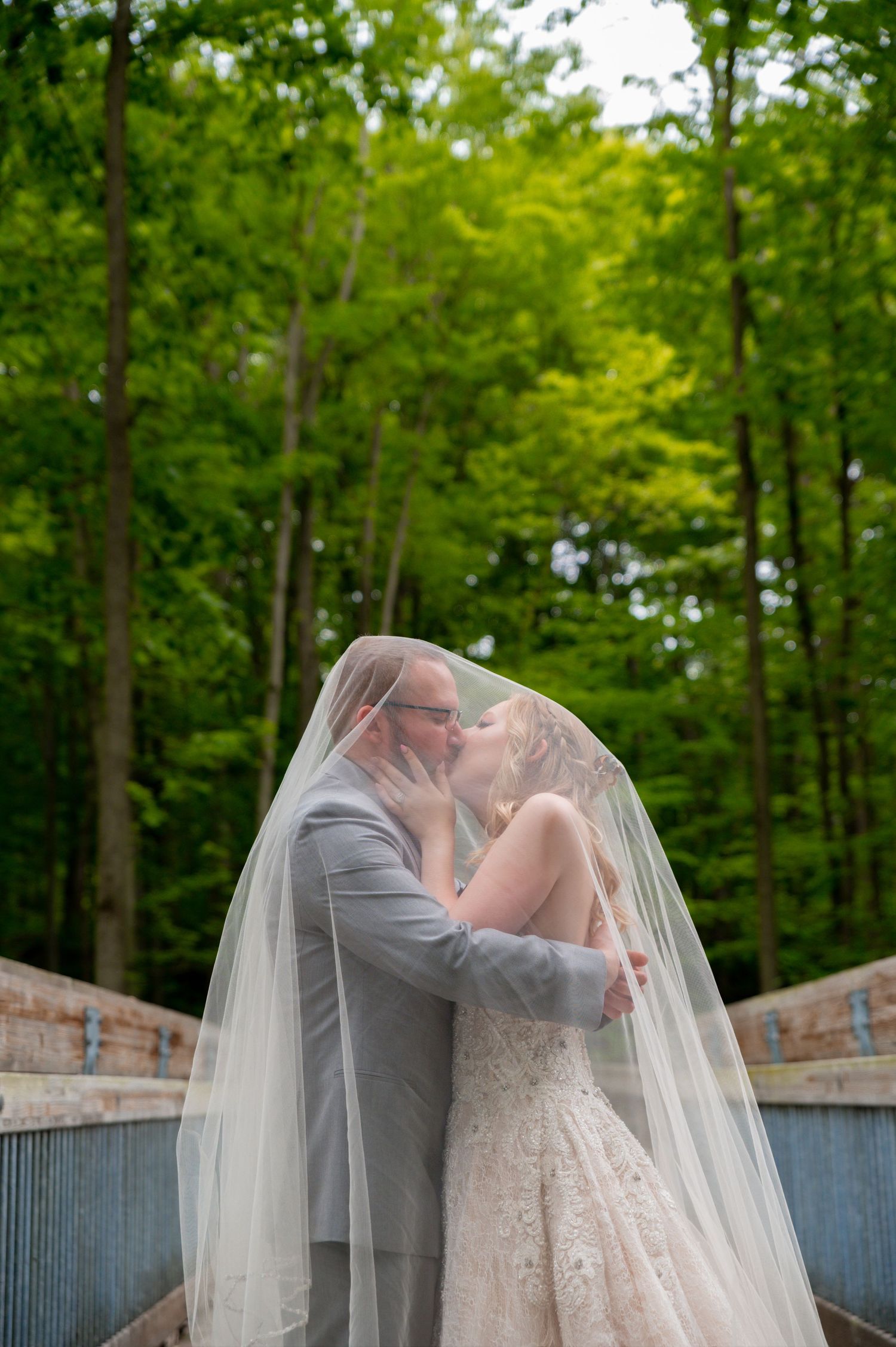 A romantic wedding couple shares a kiss under a flowing veil against a lush green forest backdrop.