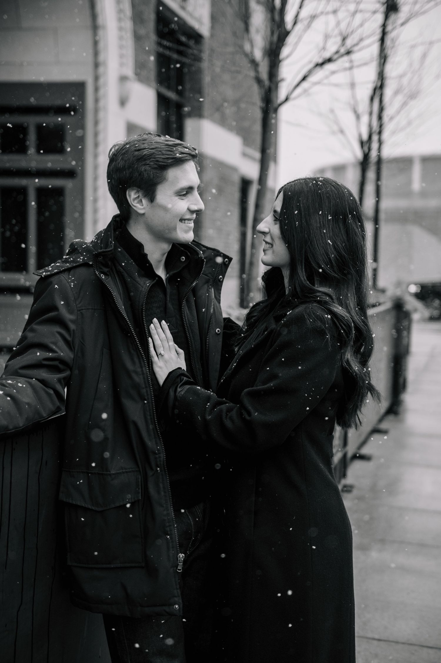 A couple shares an intimate moment in the snow wearing dark winter coats.
