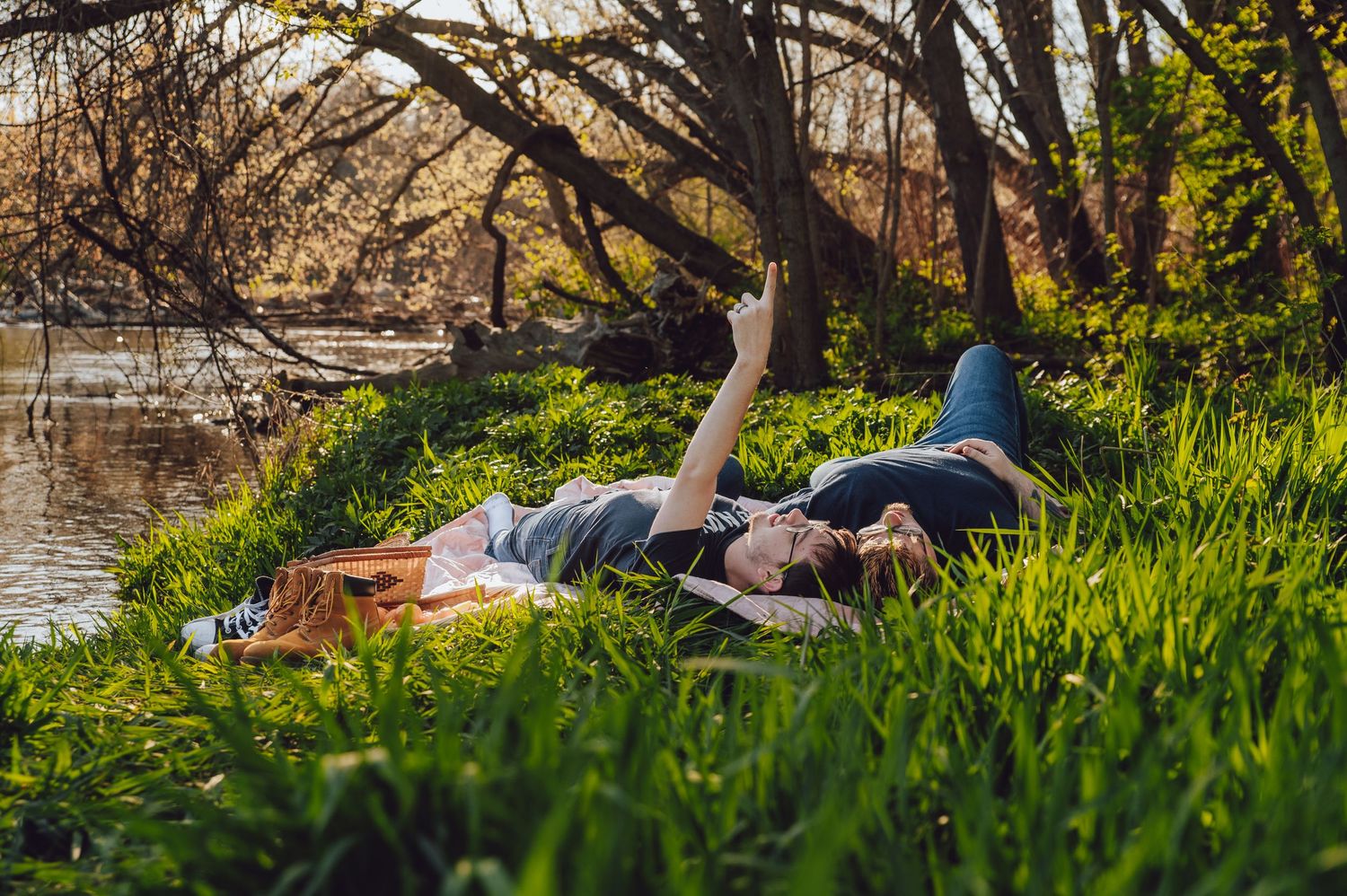 Two people lounging on grass by a riverside on a sunny afternoon.