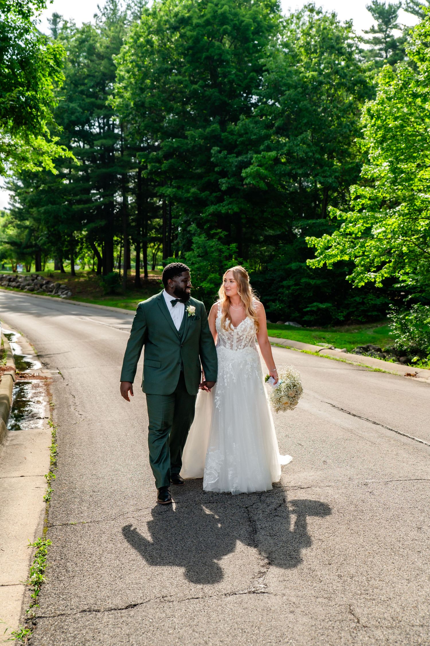 A couple in wedding attire walks down a tree-lined path enjoying a summer day.