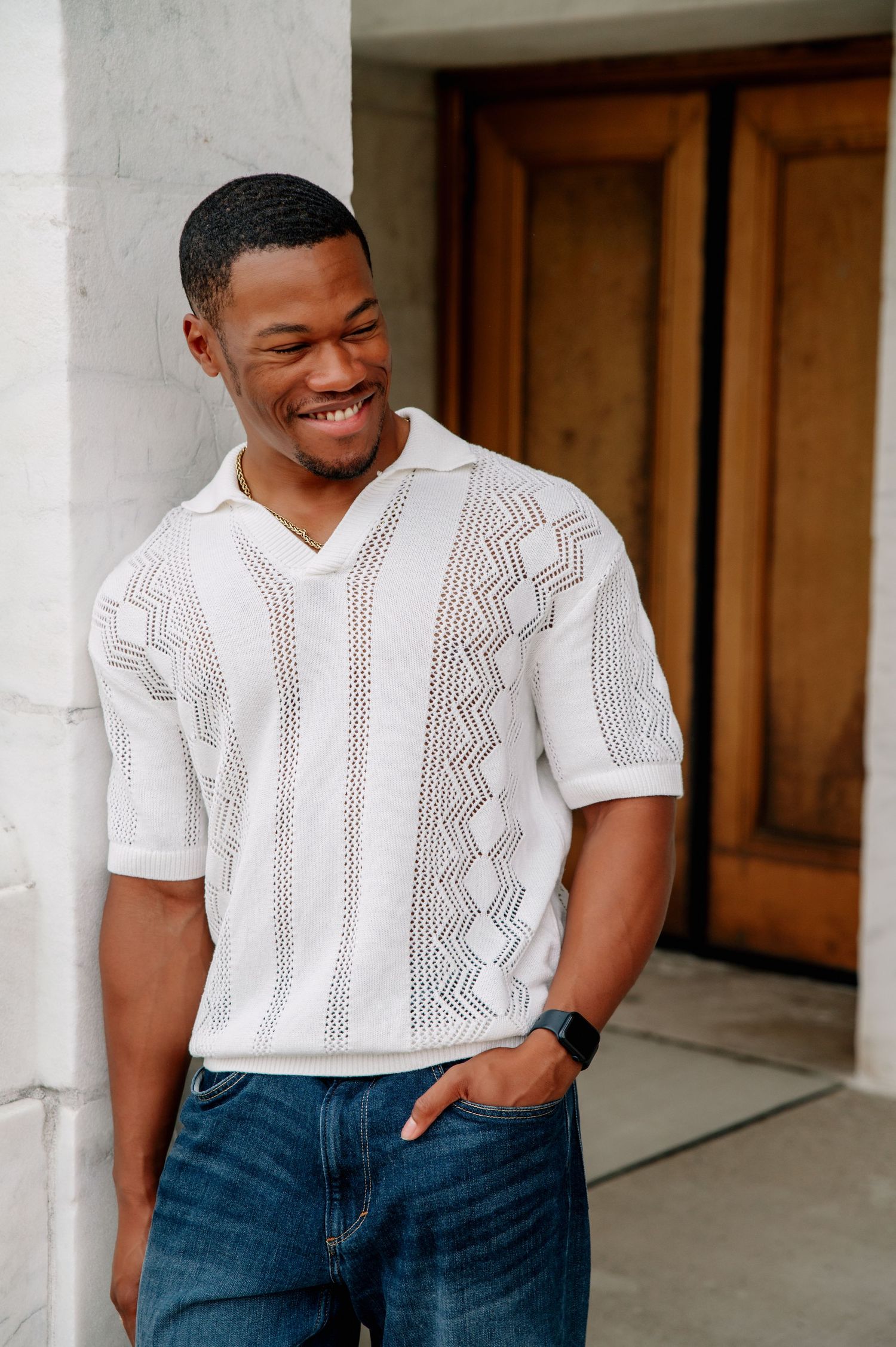 Man smiling while wearing white patterned knit shirt and jeans against marble wall.