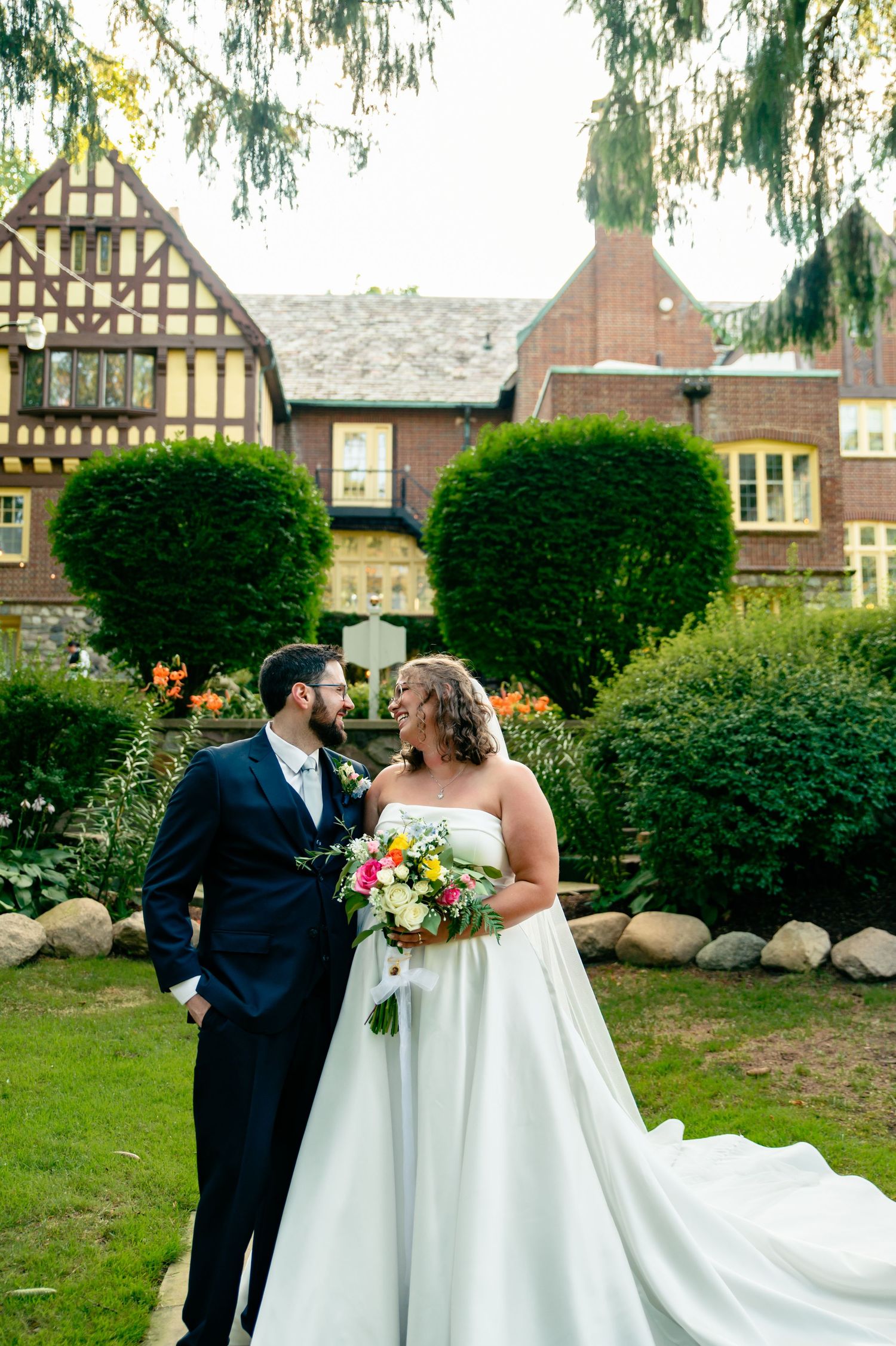 A couple in wedding attire sharing a romantic moment together in a garden setting with Tudor-style buildings in the background.