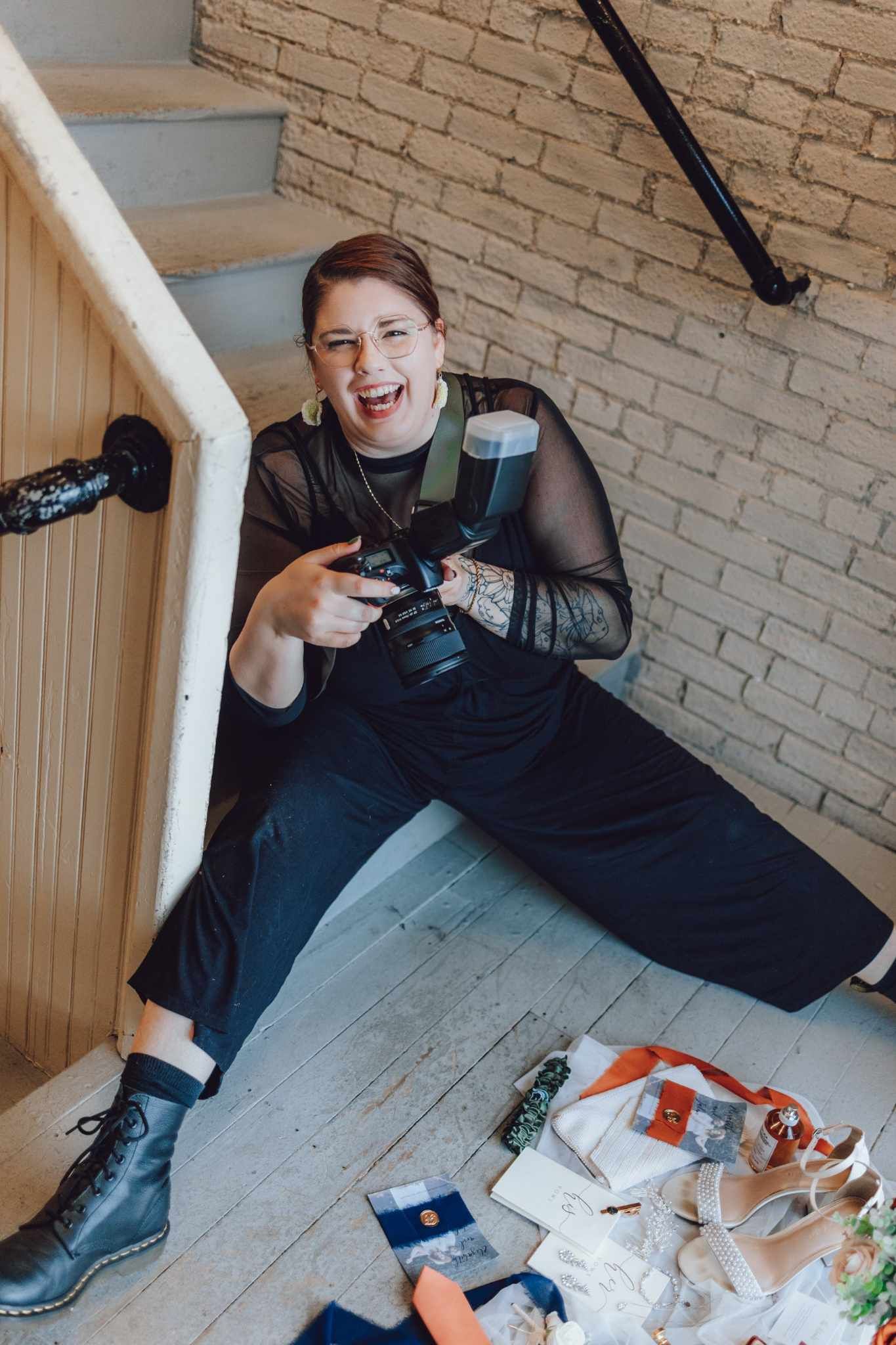 A wedding photographer sits on stairs with camera gear and bridal accessories nearby.