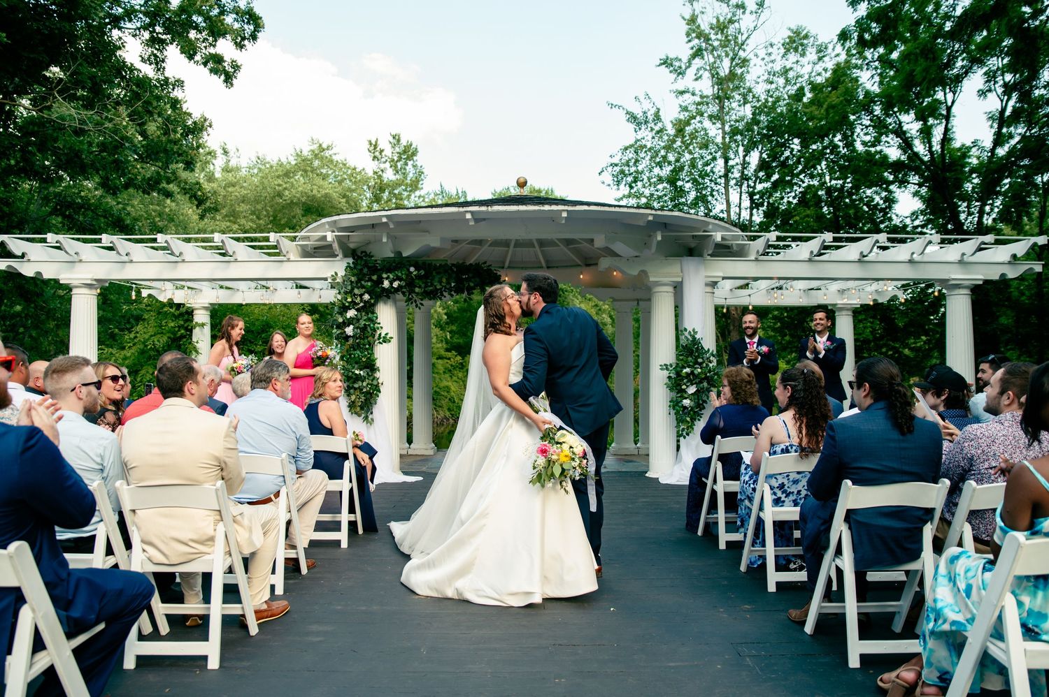 Panoramic view of an outdoor wedding reception with white folding chairs and tables set up under a pergola.