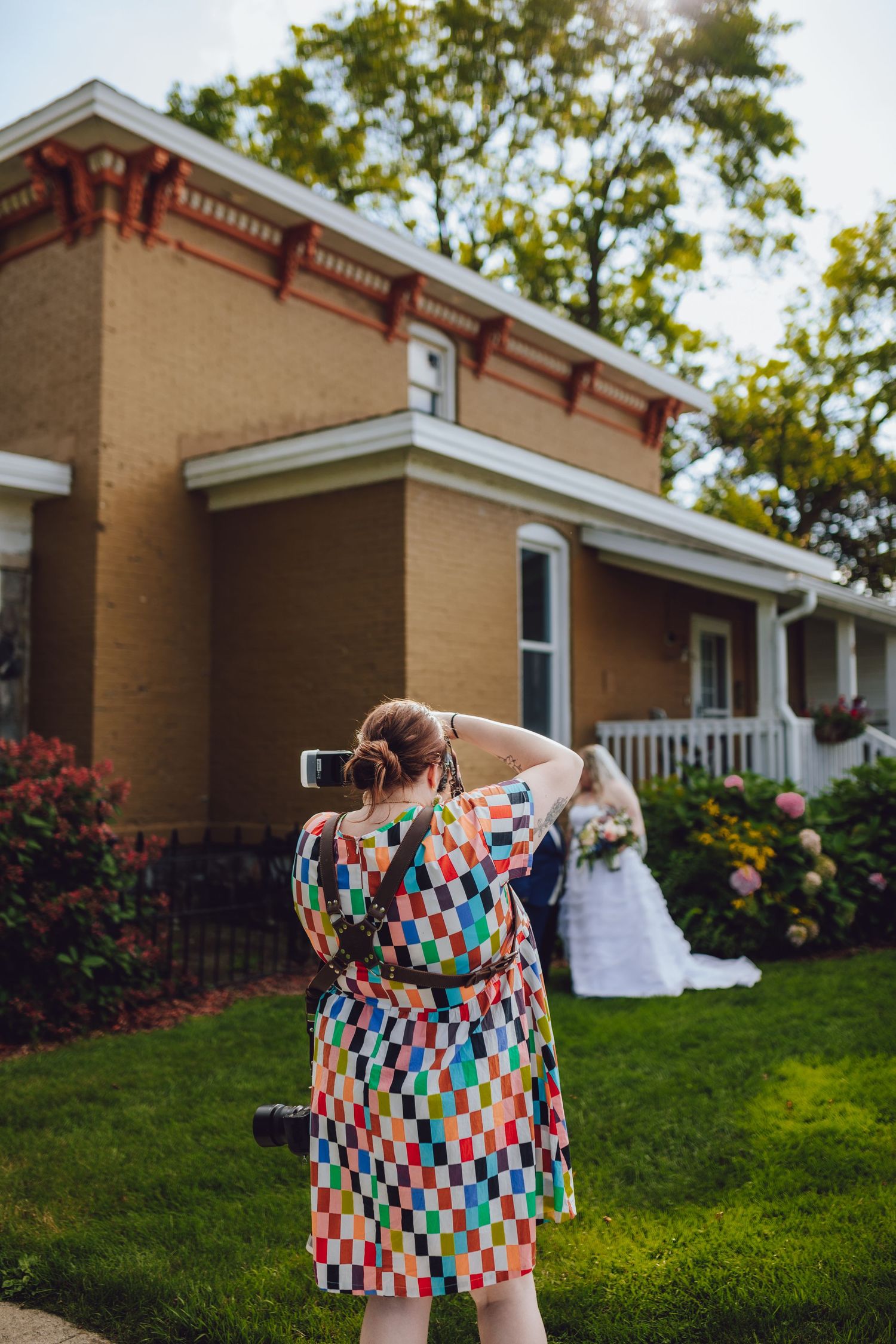 A photographer in a colorful patterned dress captures a wedding moment in front of a historic brick house with a porch.