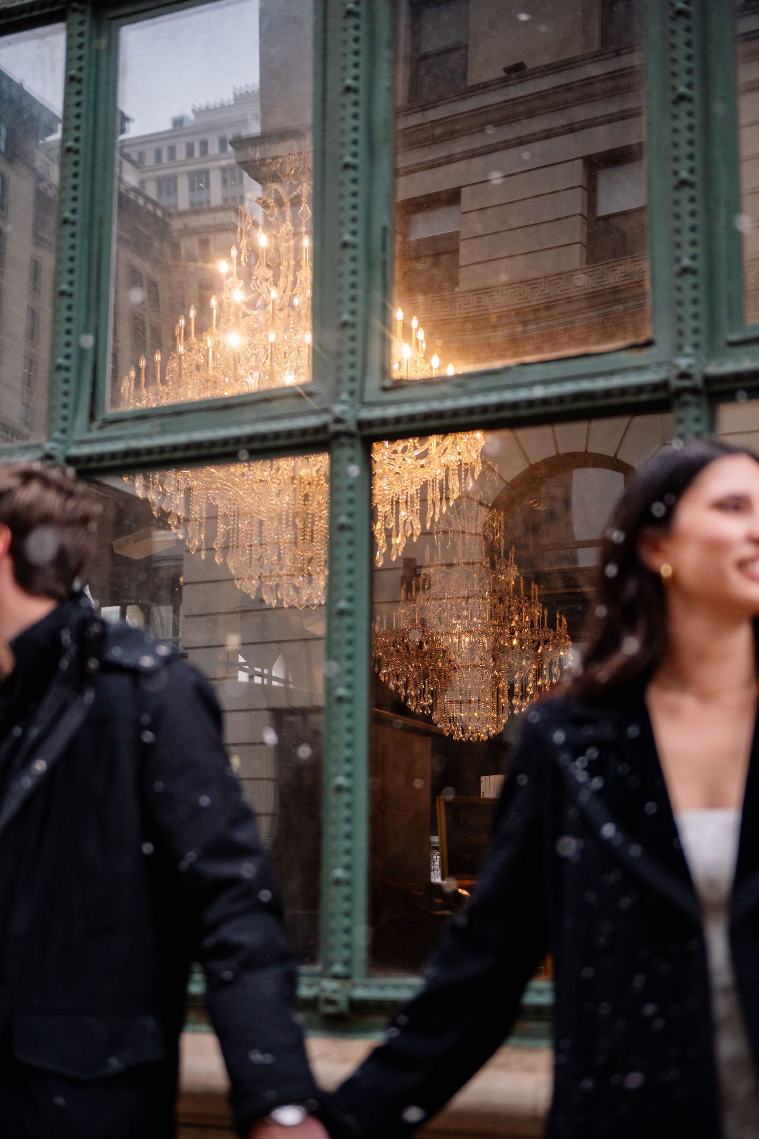 A couple walks past an illuminated window display with a chandelier reflection on a snowy evening.