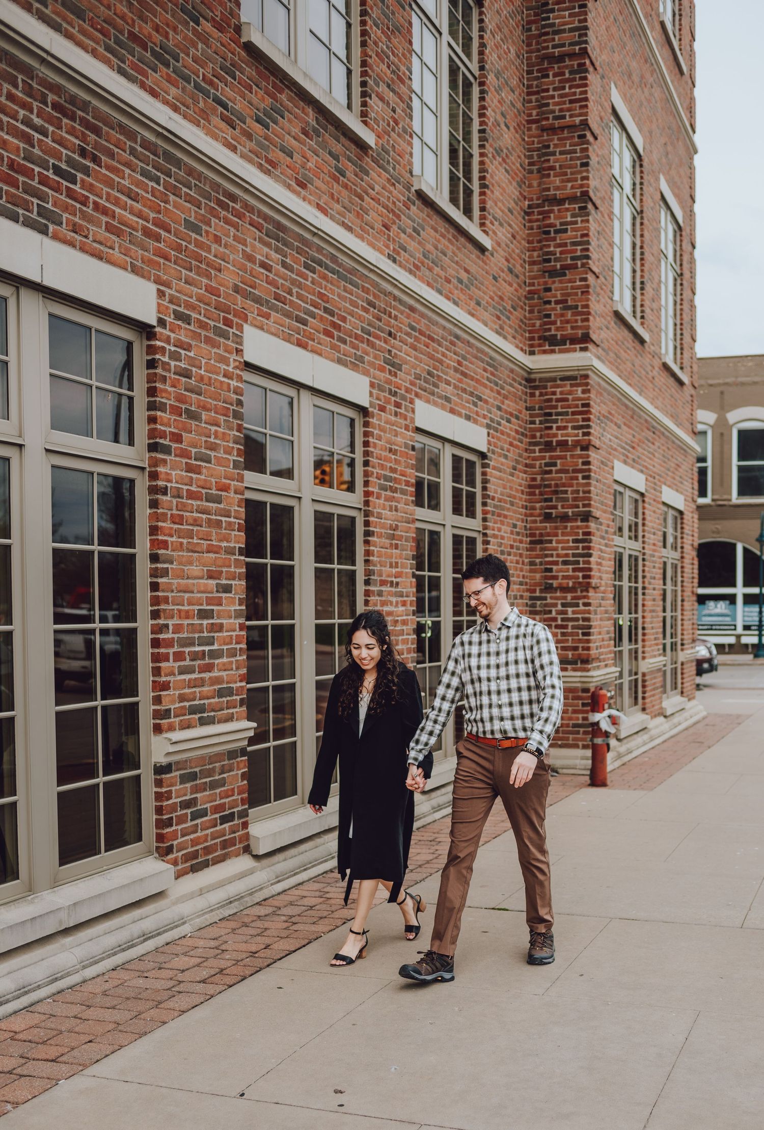 Couple walking along historic brick building with large windows on urban sidewalk.