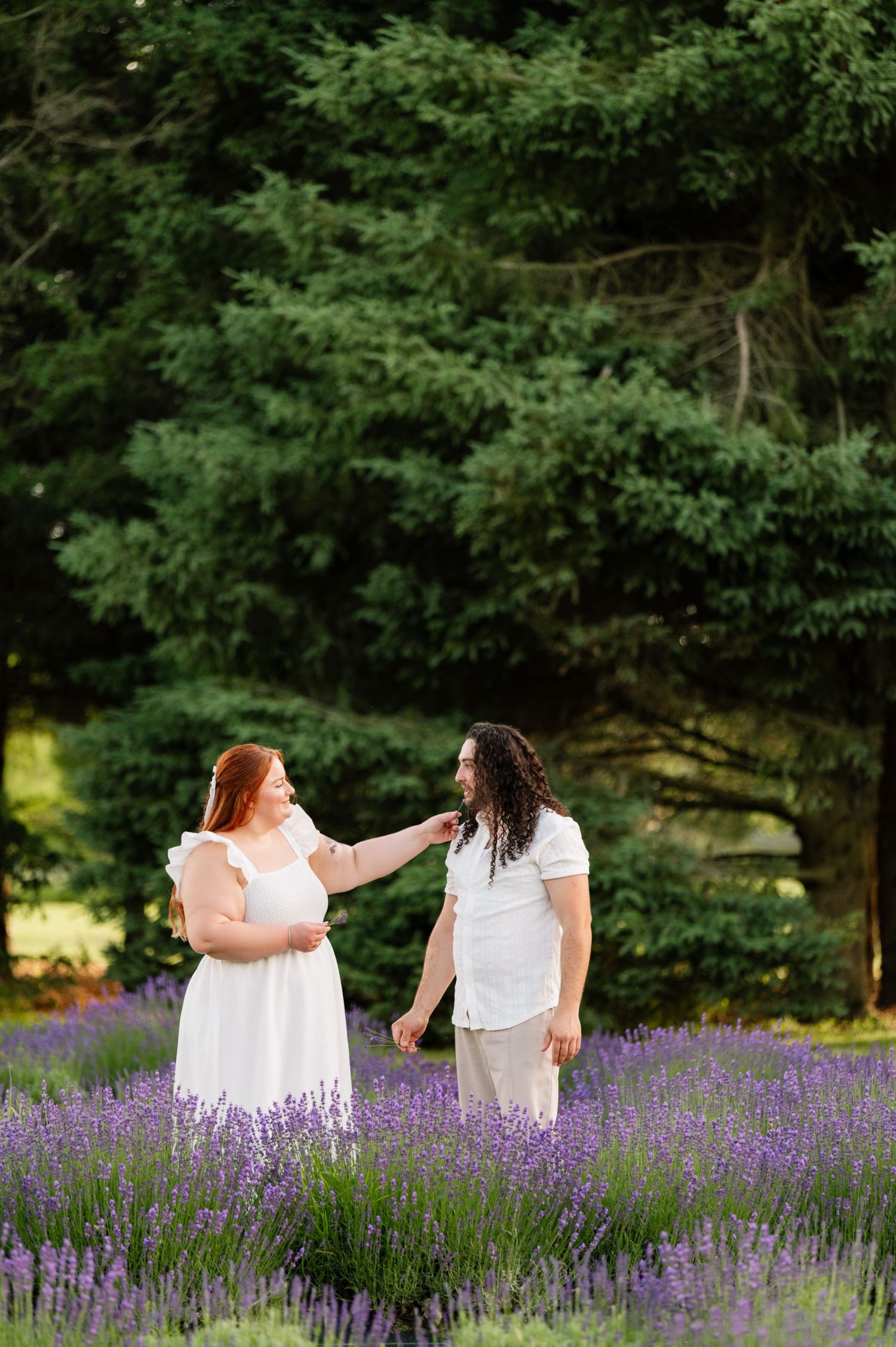 Two people share a tender moment in a blooming lavender field with trees in the background.