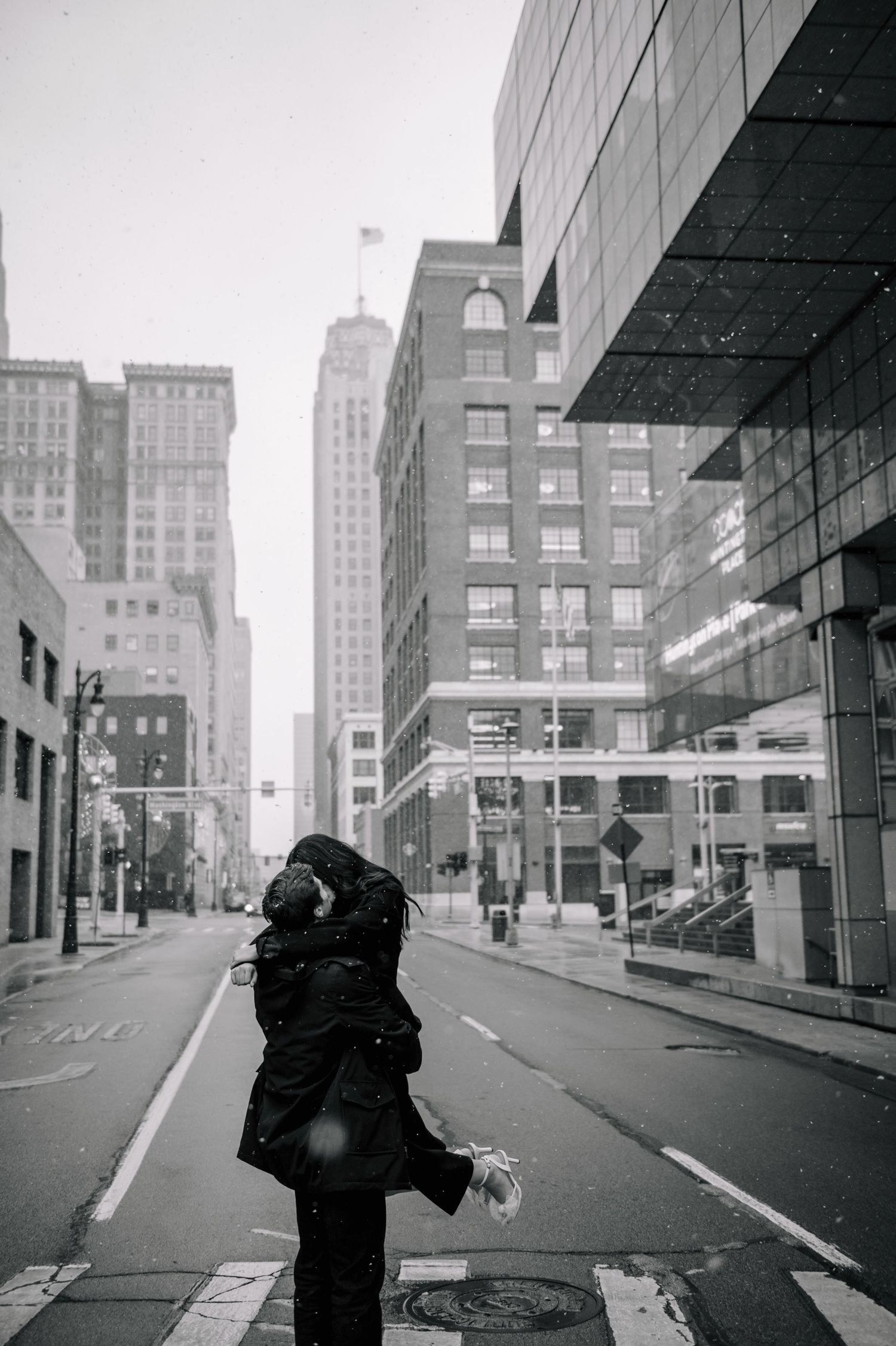 A couple embraces in a black and white cityscape on an empty street with tall buildings.
