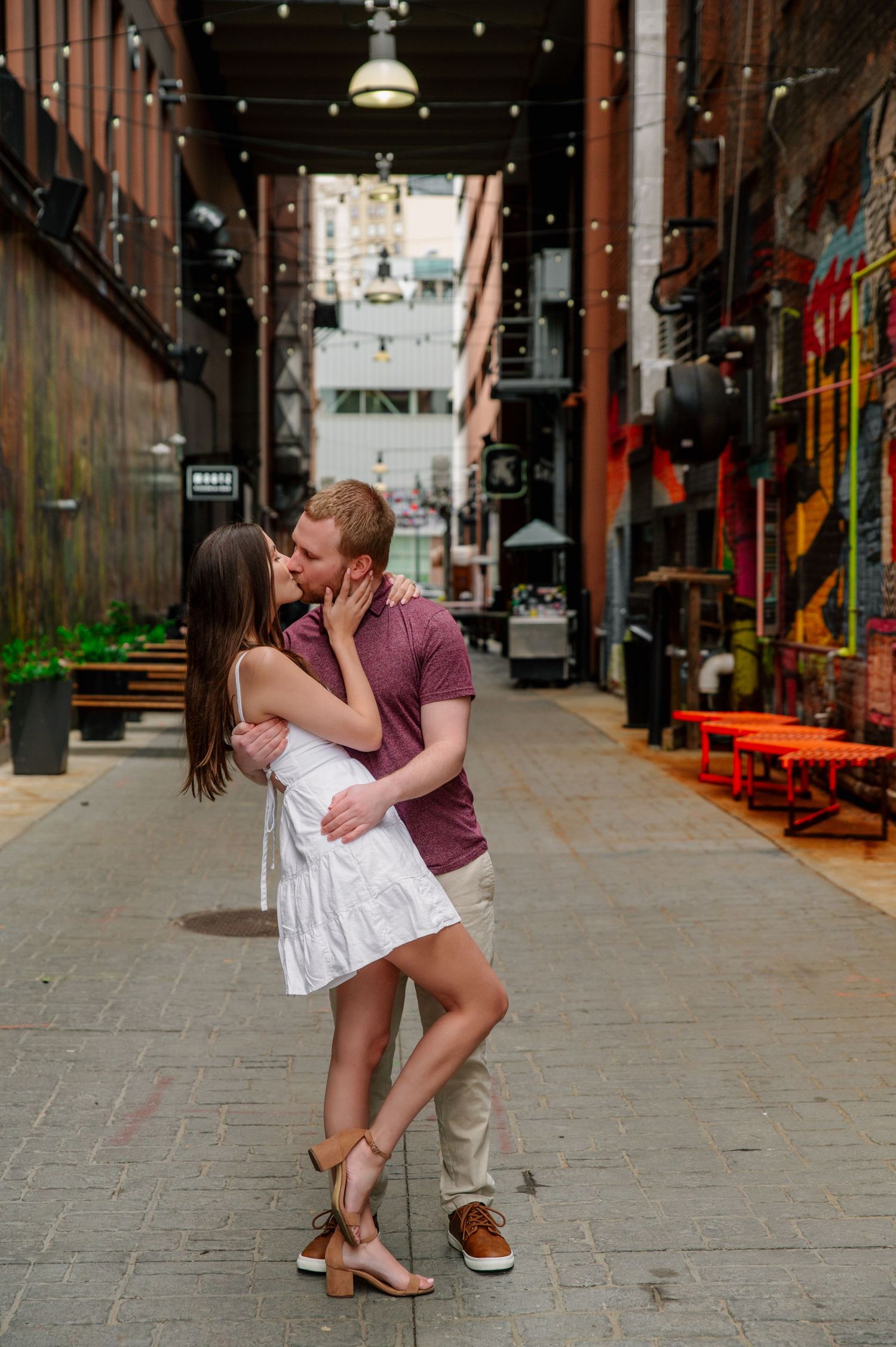 A couple shares a romantic kiss in an urban alley decorated with string lights and graffiti.