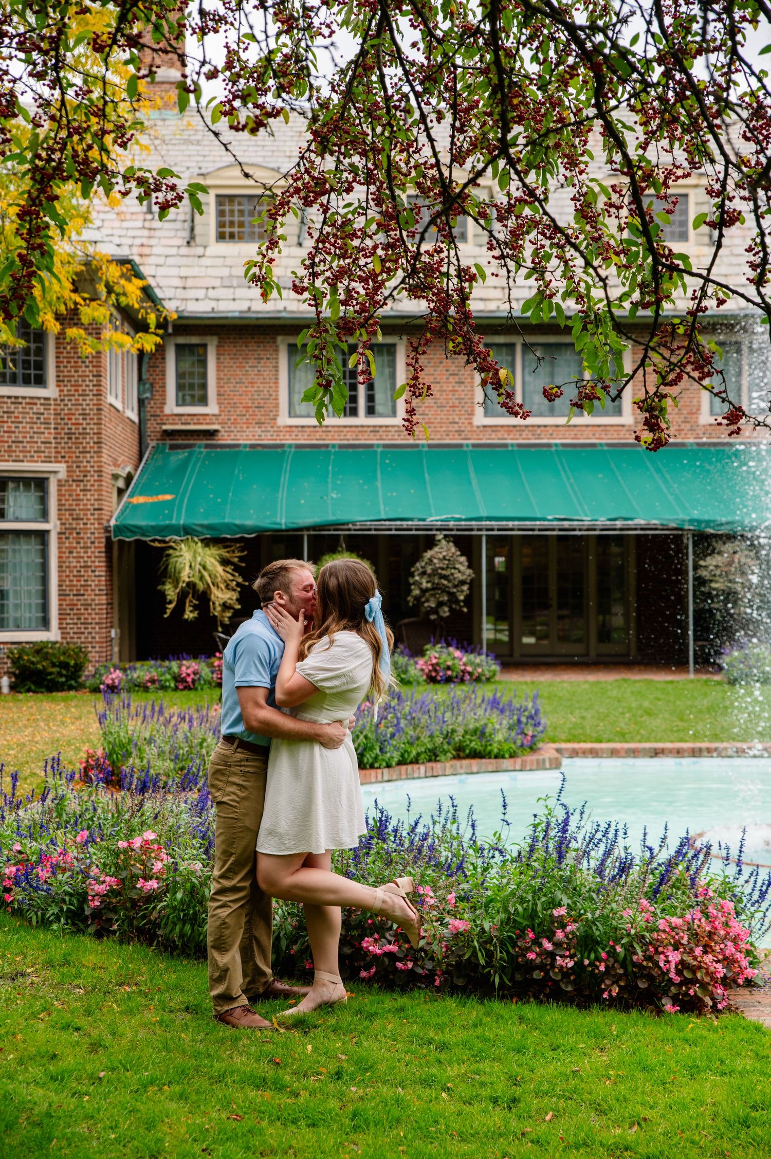 Couple shares romantic kiss in front of brick building with blooming garden.