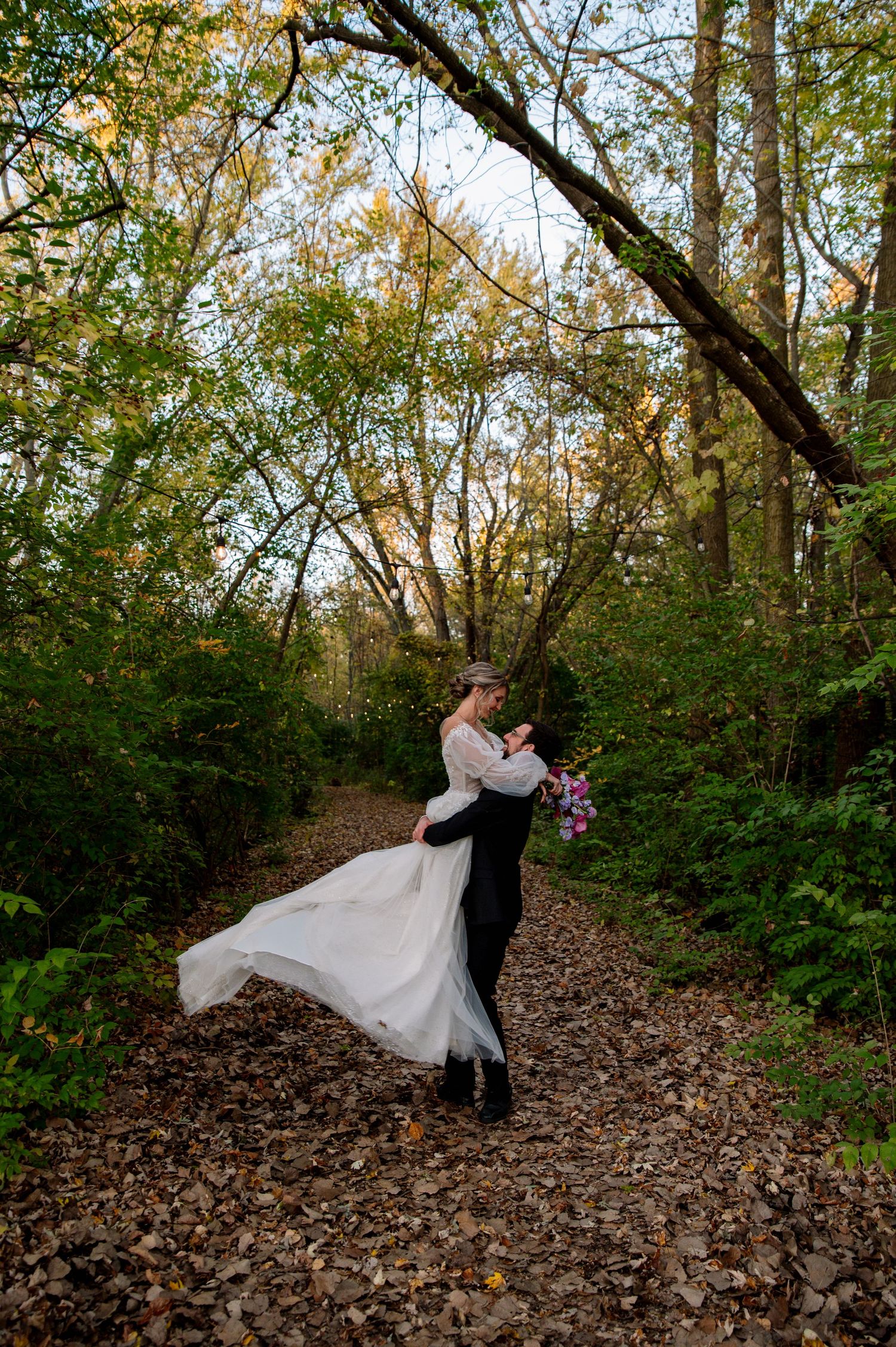 A romantic couple dancing and embracing on a path covered in autumn leaves surrounded by green forest trees.