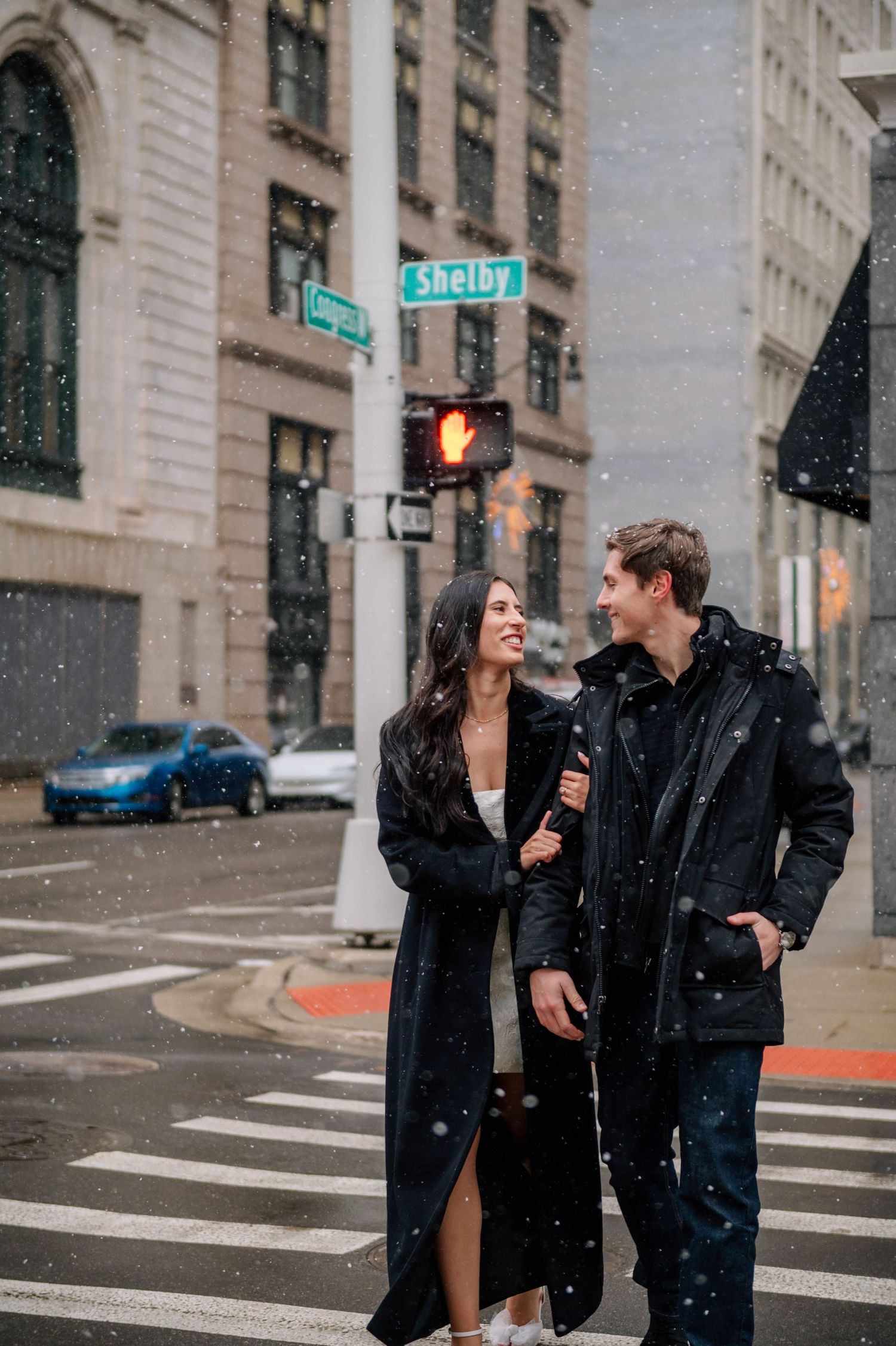 A couple walks together during snowfall at a city intersection with traffic lights.