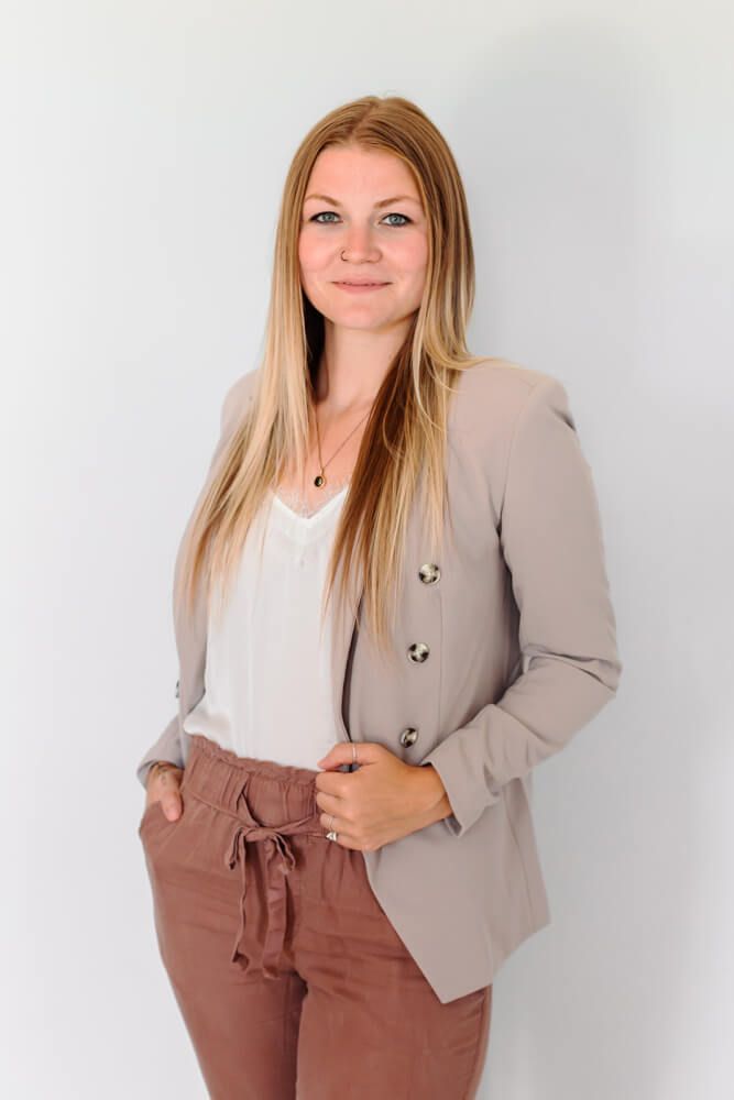 A professional woman in beige blazer and white top poses against white background.