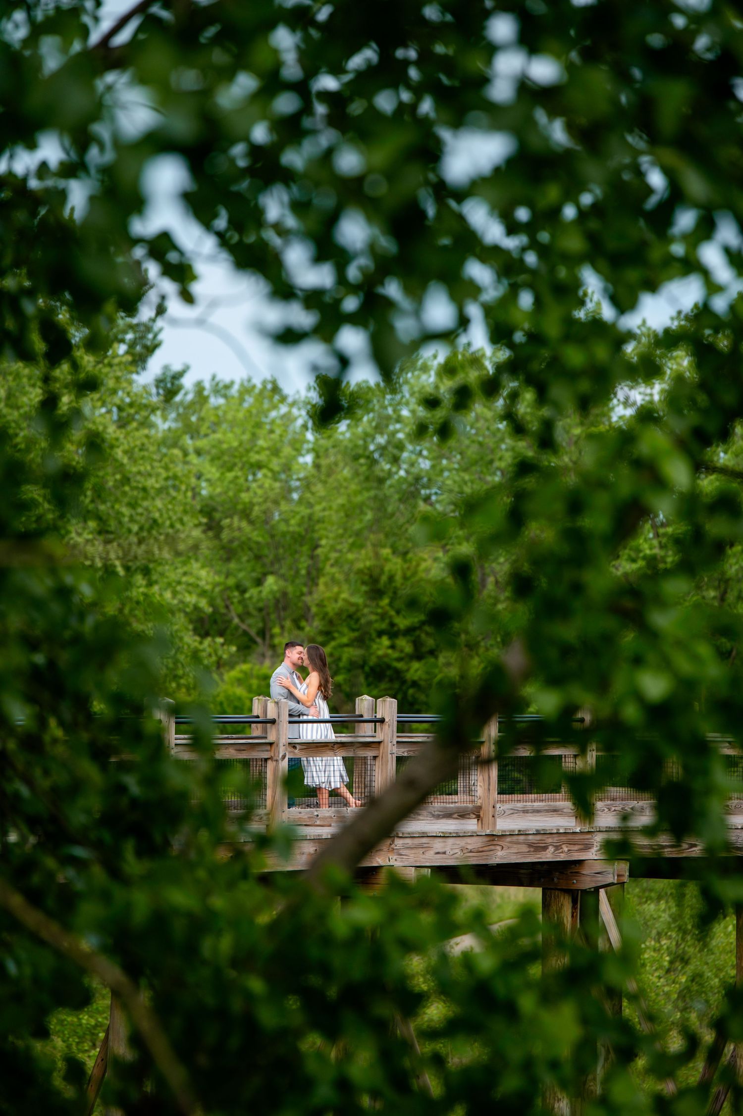 A couple embraces on a wooden boardwalk surrounded by lush green foliage on a summer day.