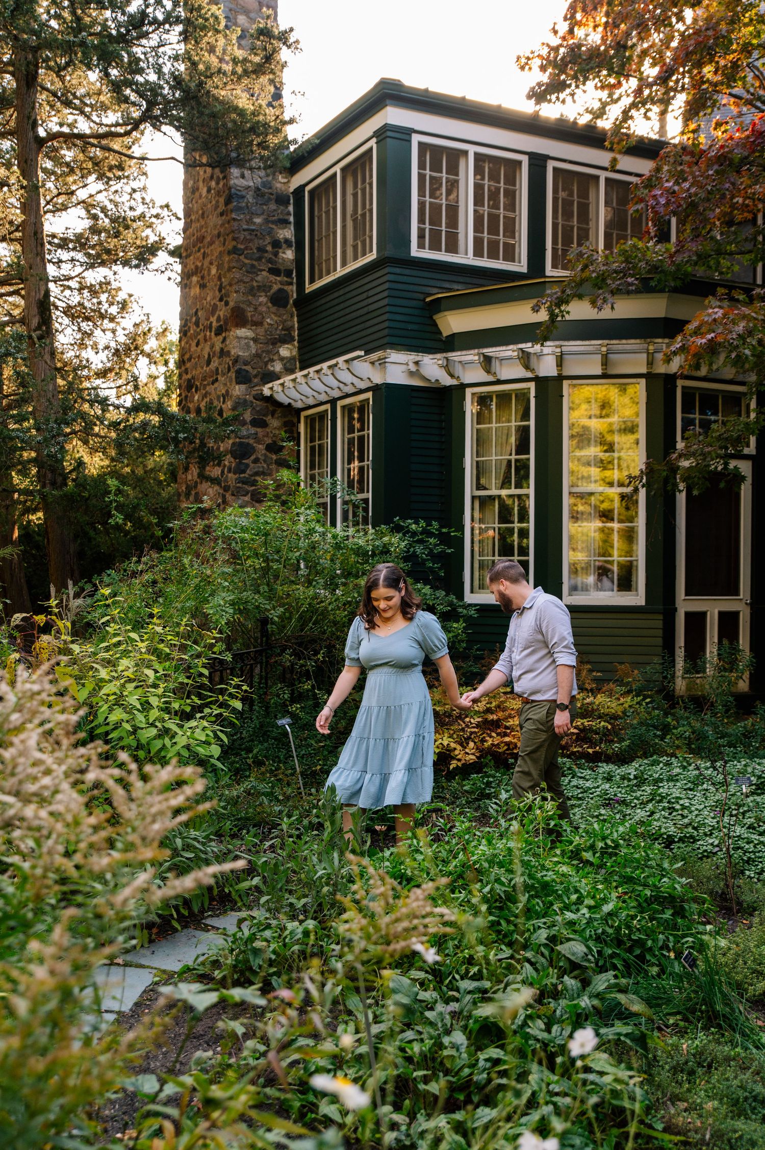 Couple exploring garden path around charming dark green Victorian house with bay windows in golden afternoon light.