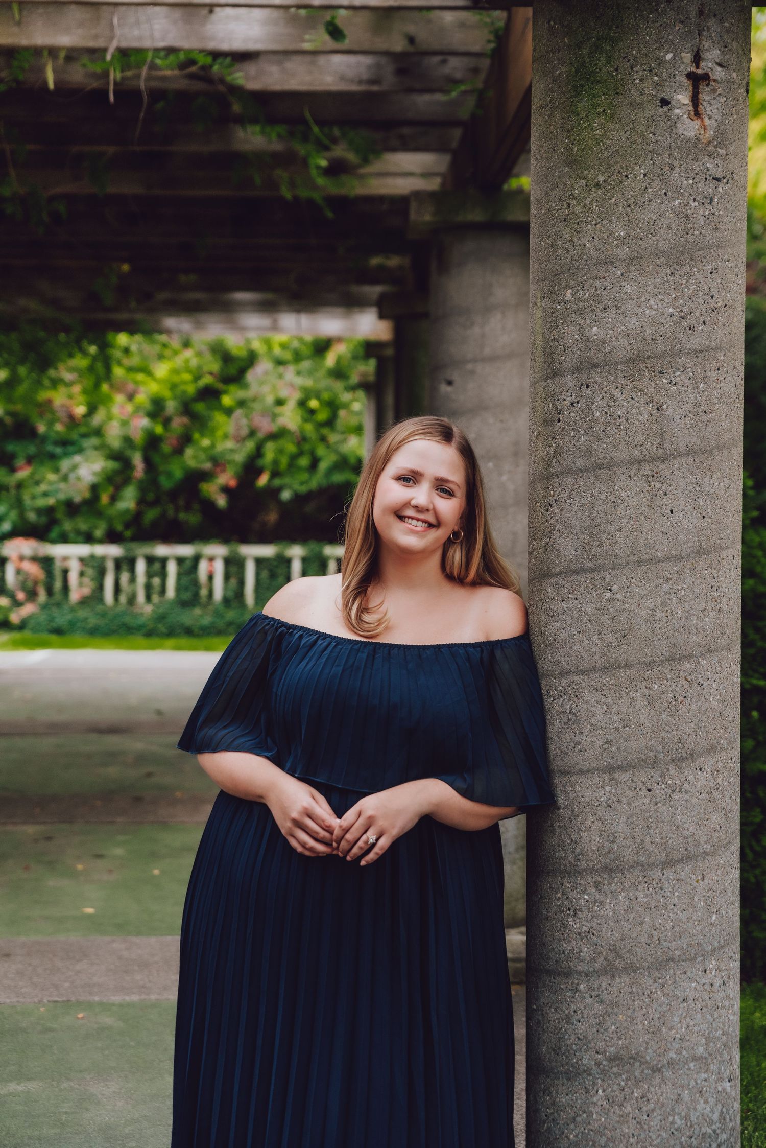 Person in navy off-shoulder dress smiling against concrete pillar in garden setting.