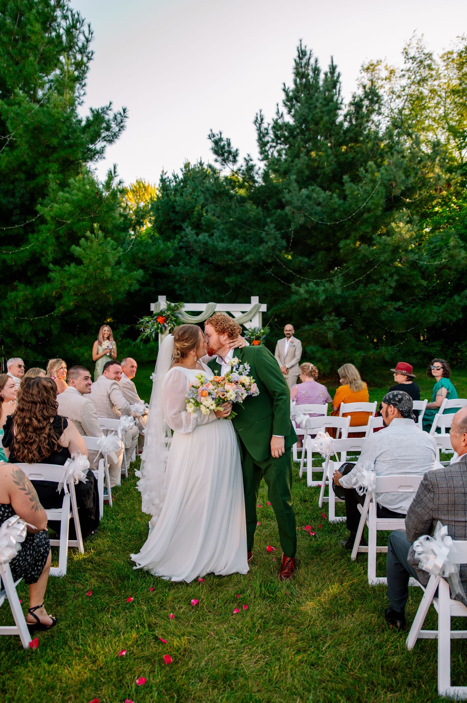 An outdoor wedding ceremony in a pine forest with white chairs and gathered guests.