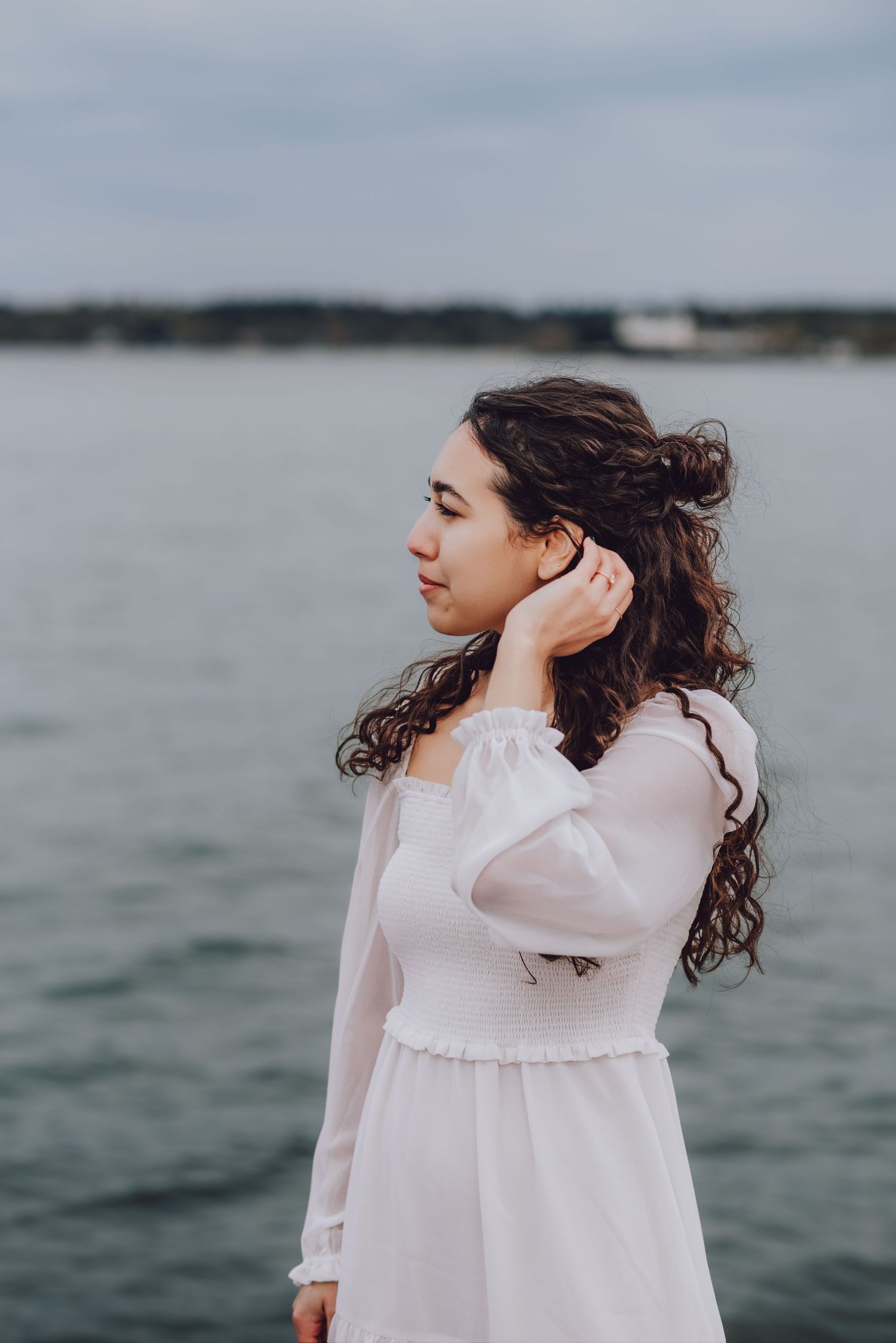 A person in a white dress stands near the water on a cloudy day.