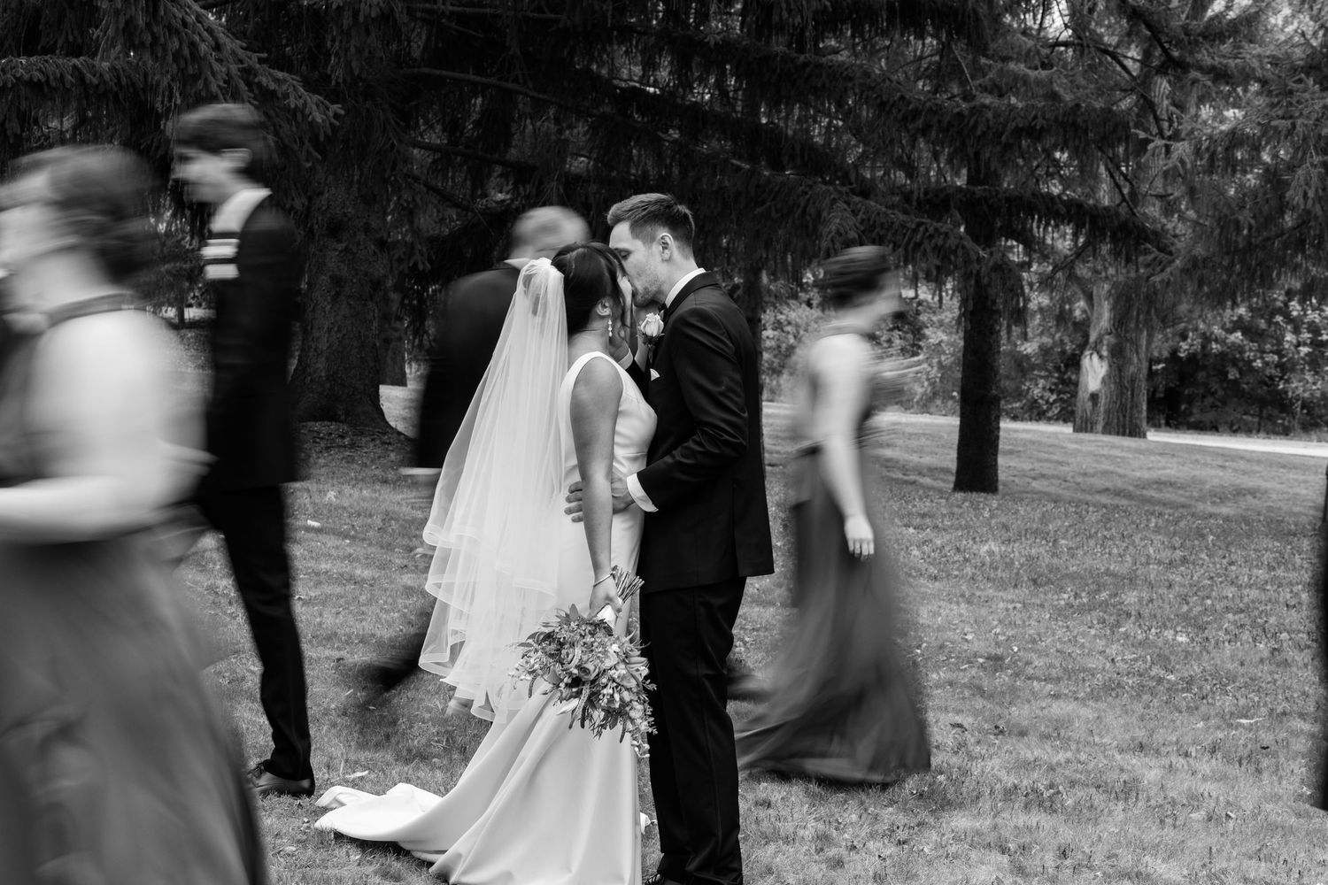 Black and white wedding photograph showing couple during outdoor ceremony with blurred motion of guests moving around them.