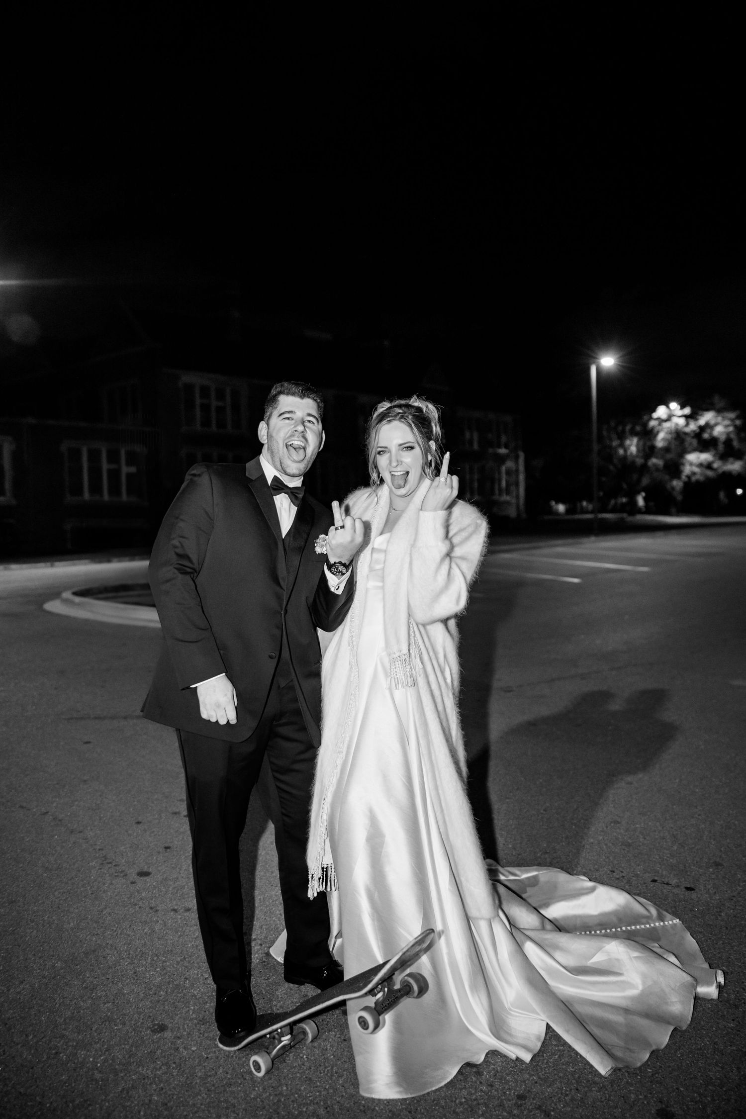 A couple in formal attire playfully pose with skateboards at night in black and white photography.