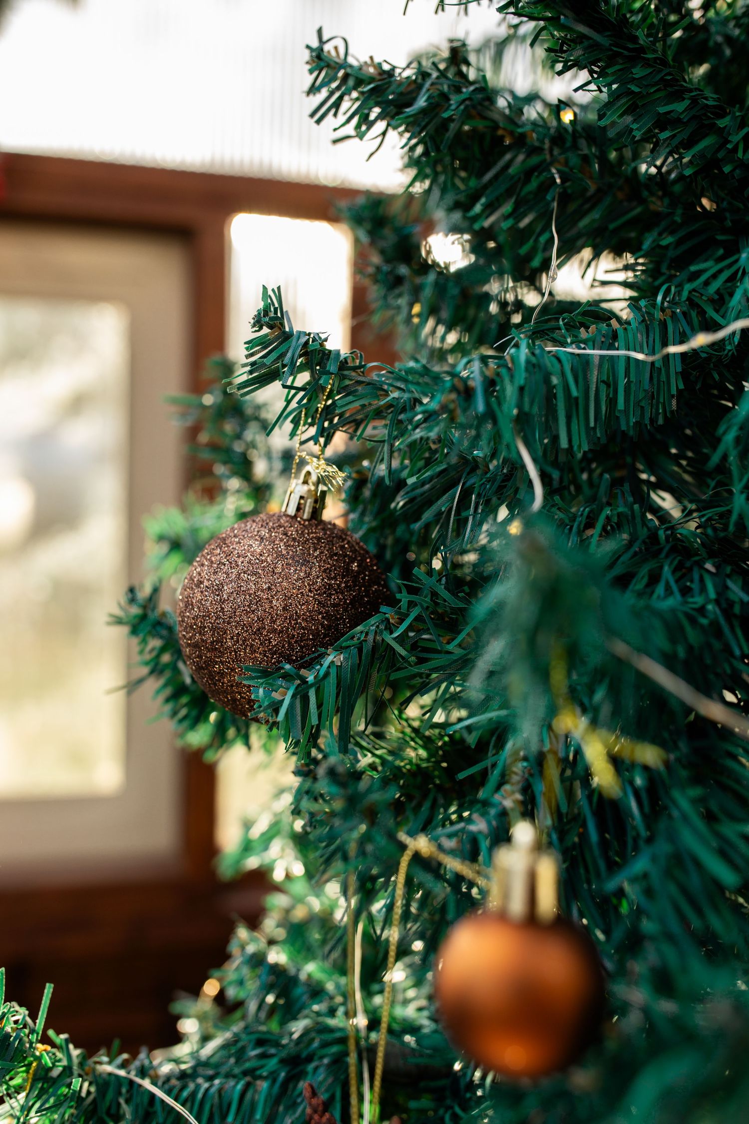 Close-up of brown glittery Christmas ornaments hanging on green artificial tree branches with soft window light.