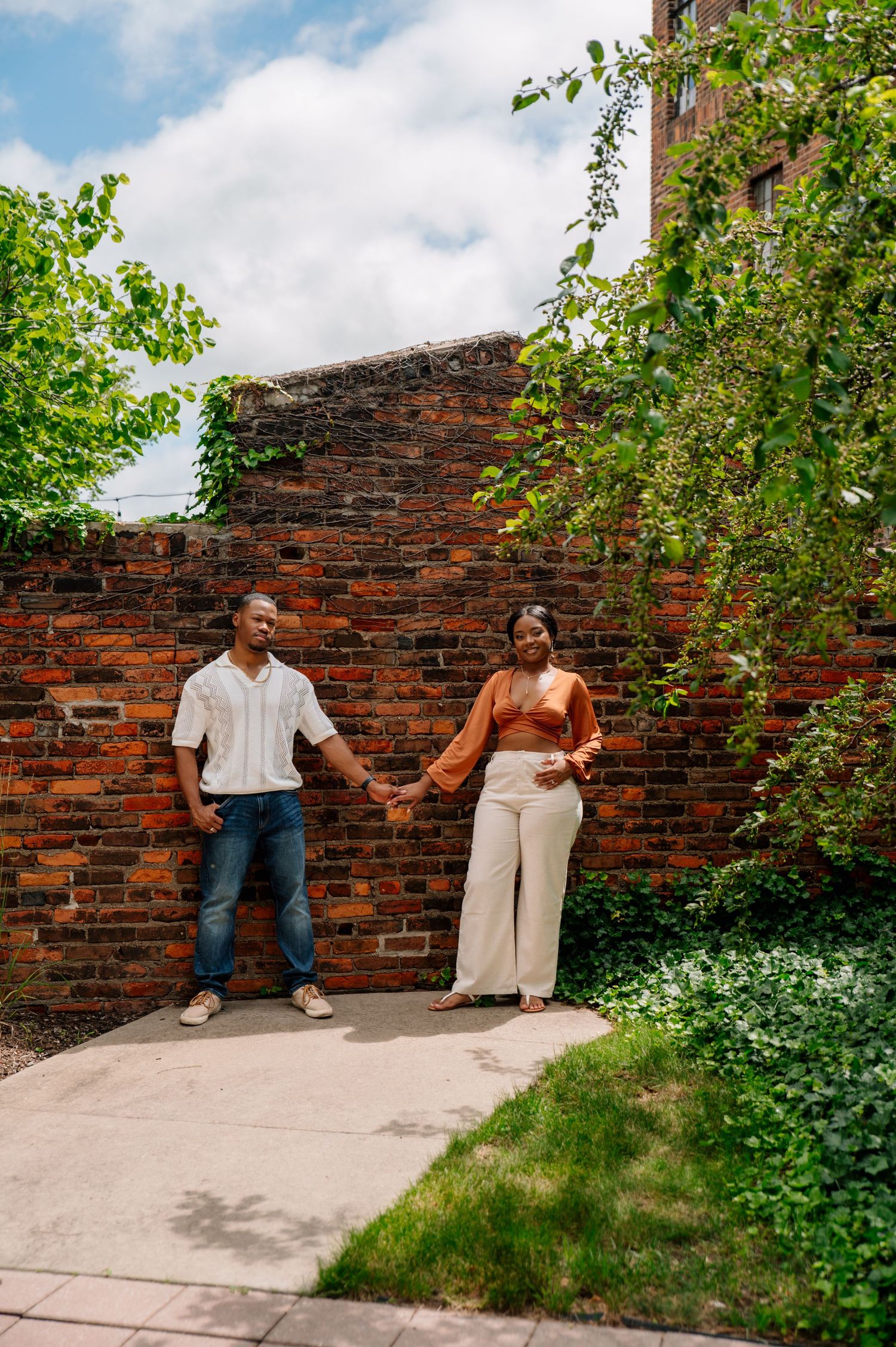 Stylish couple posing against brick wall with greenery in urban setting.