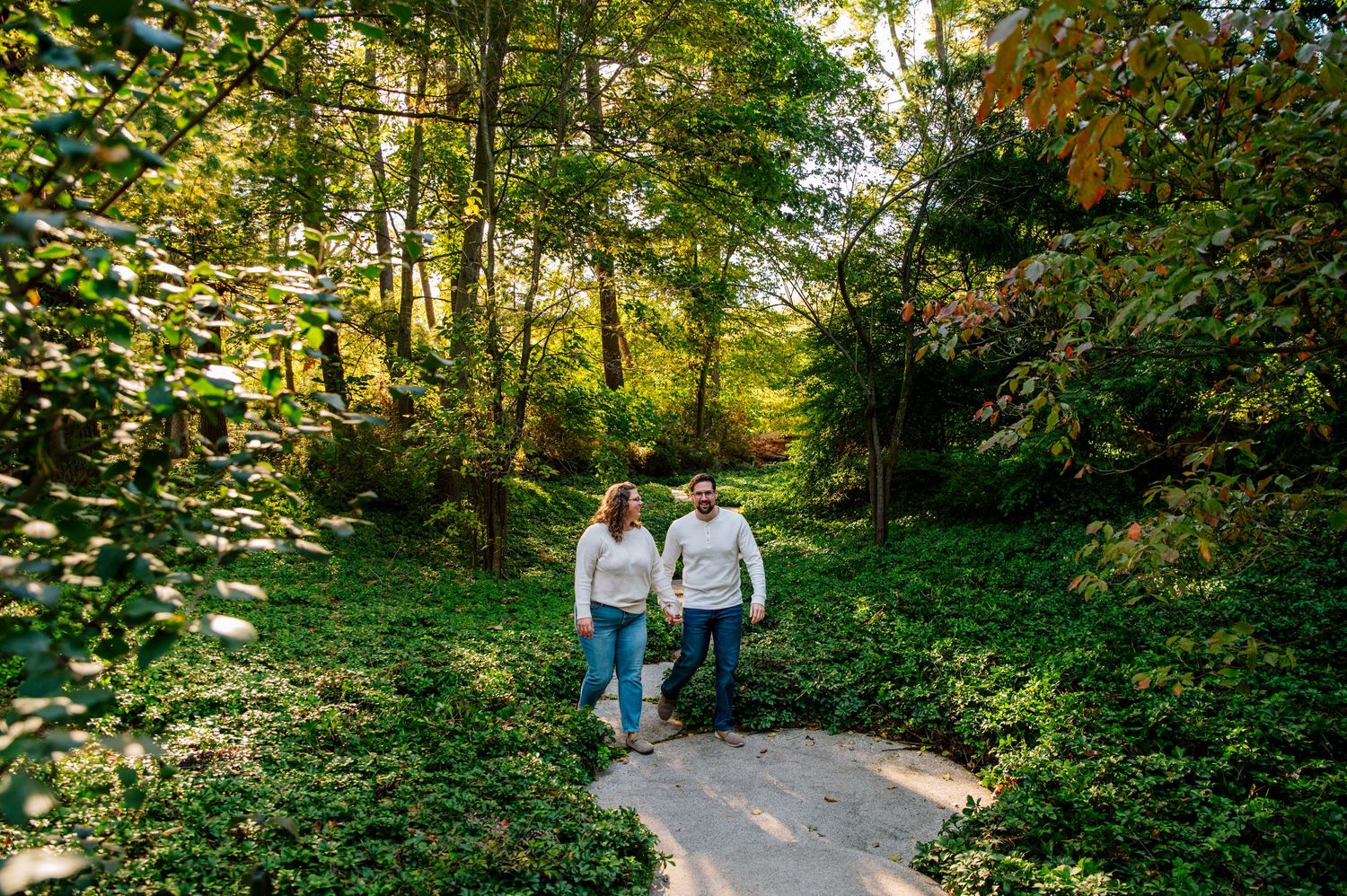 A couple walks hand in hand along a sunlit path through lush green woods.