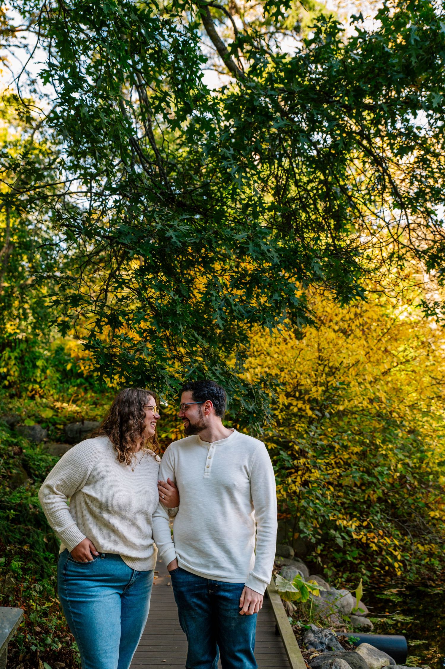 A couple in coordinating white sweaters share a tender moment on a wooded path.