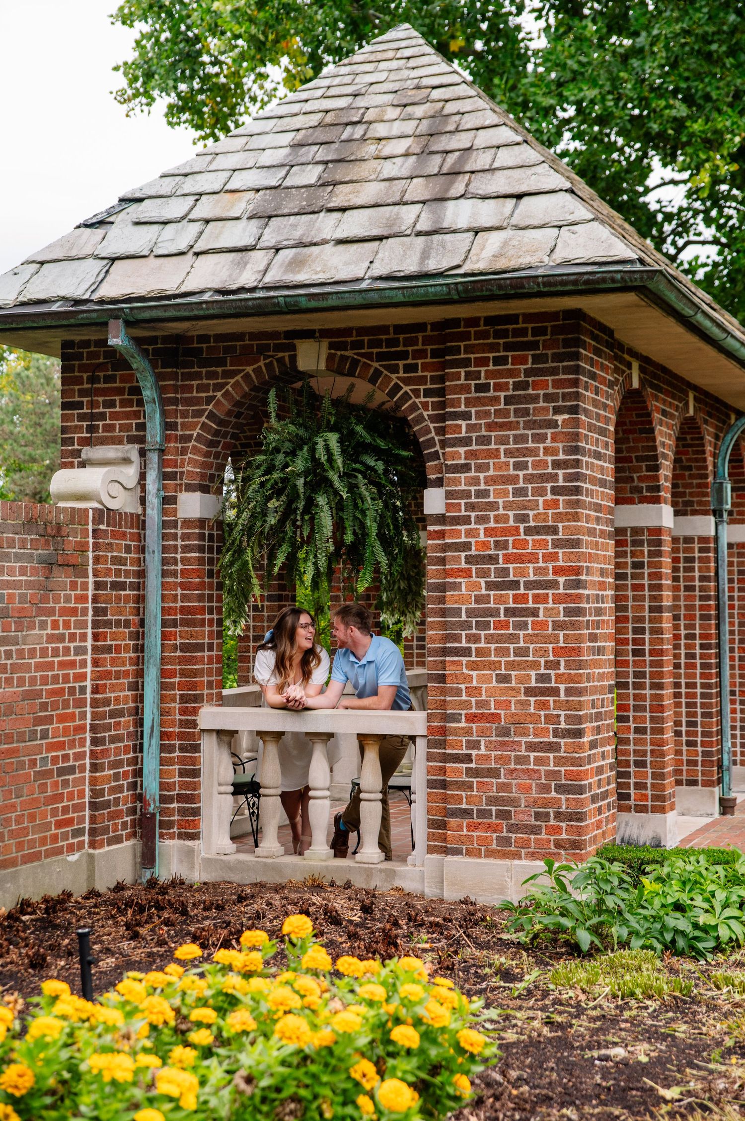 Couple leans on brick gazebo railing surrounded by yellow flowers and hanging plants.