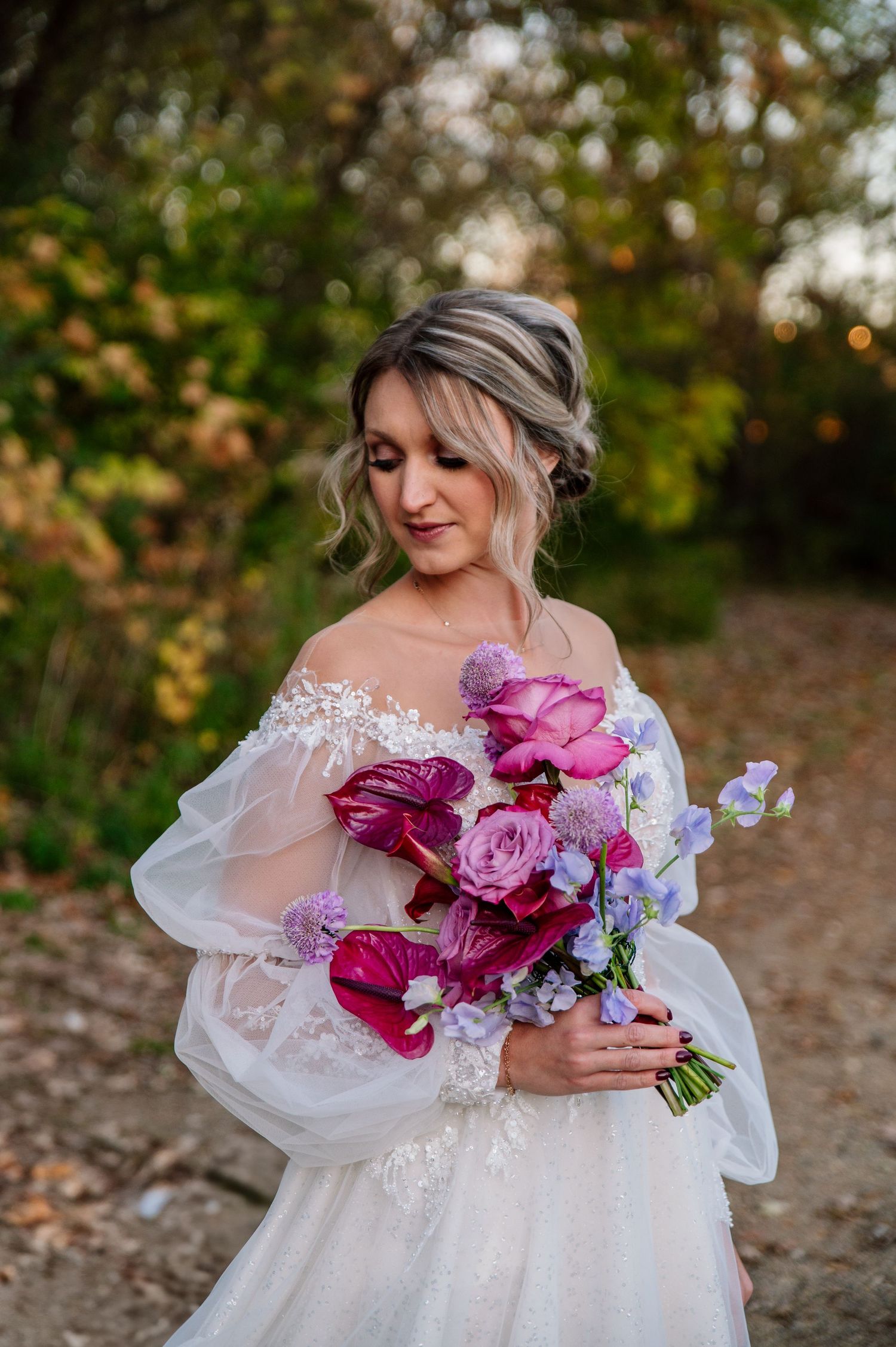 A bride in a delicate white lace gown holds a vibrant bouquet of pink and magenta flowers in an outdoor autumn setting.
