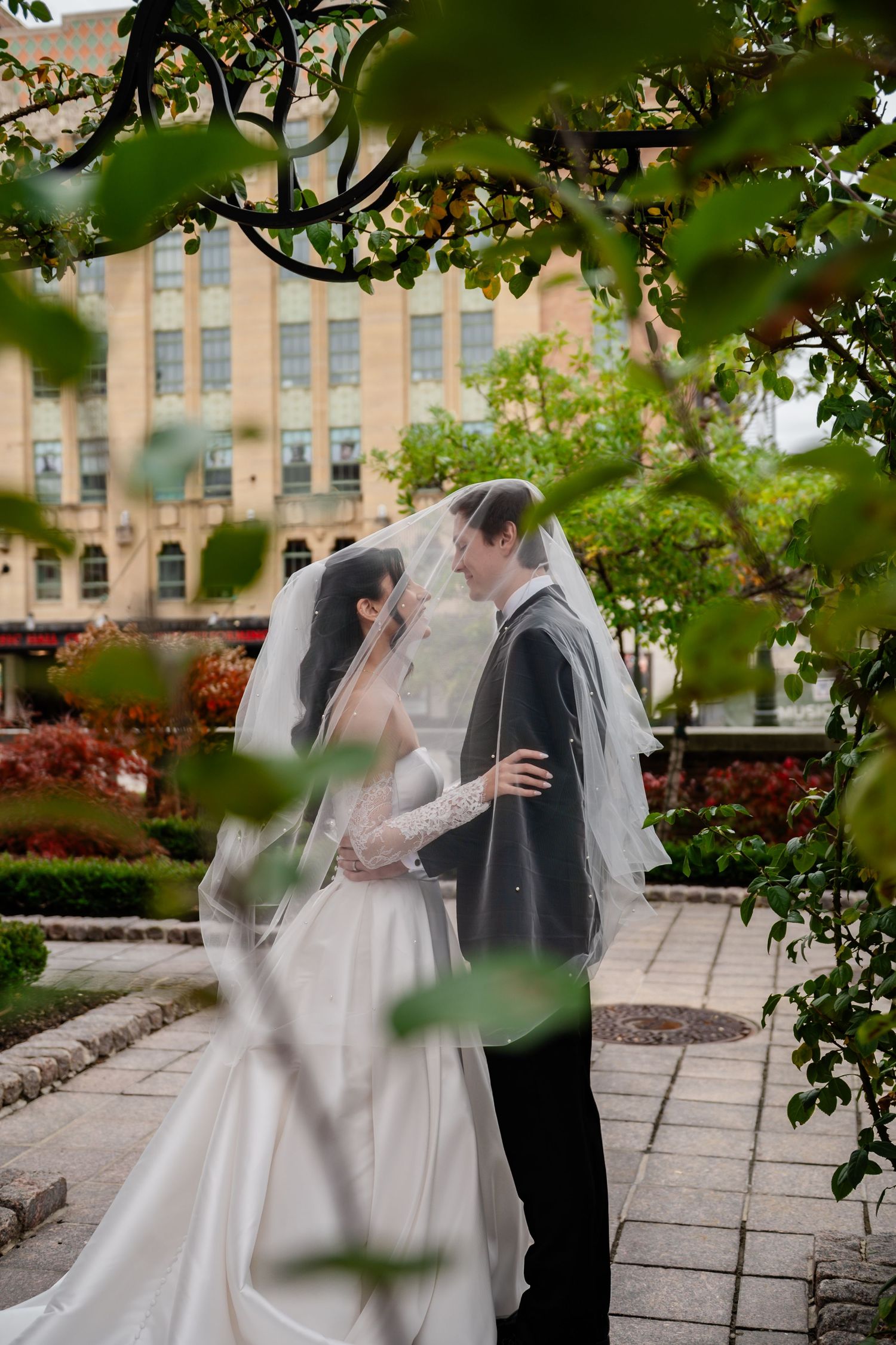 A romantic wedding couple shares an intimate moment in a garden setting with brick buildings in the background.