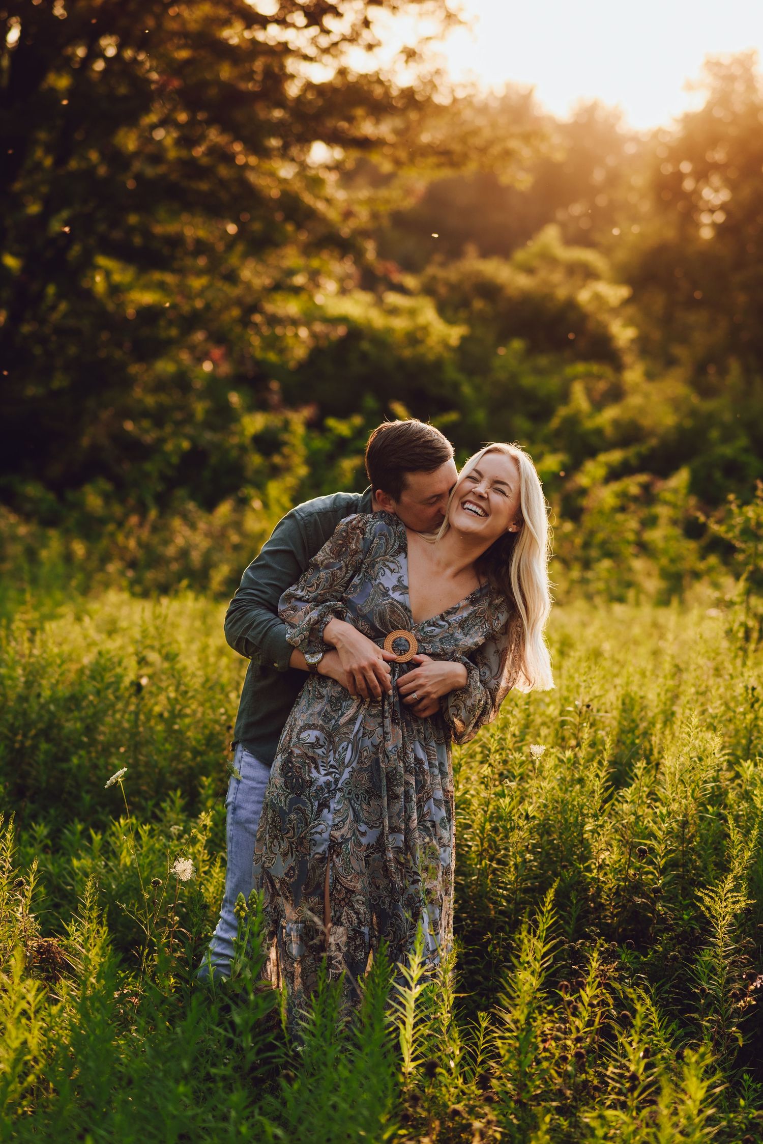 Couple sharing intimate moment in golden sunlit meadow with wildflowers and trees in background.