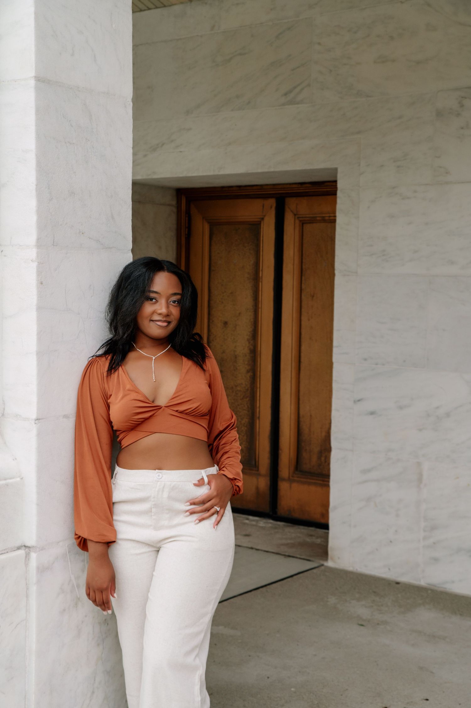 Woman in rust-colored top and white pants posing confidently against marble wall.