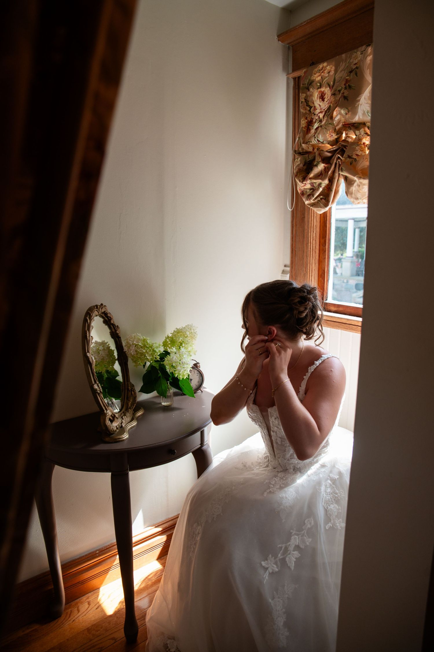 A person in a white wedding dress sits by a window in a dimly lit room with a small side table and floral arrangement.
