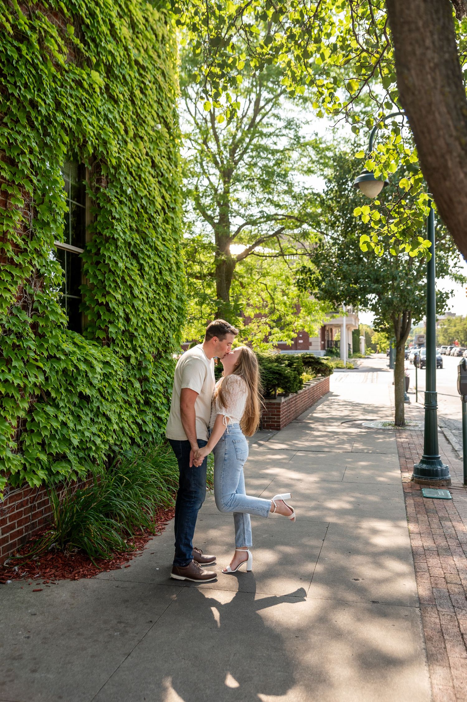 A couple shares an intimate moment on a sidewalk next to an ivy-covered wall.