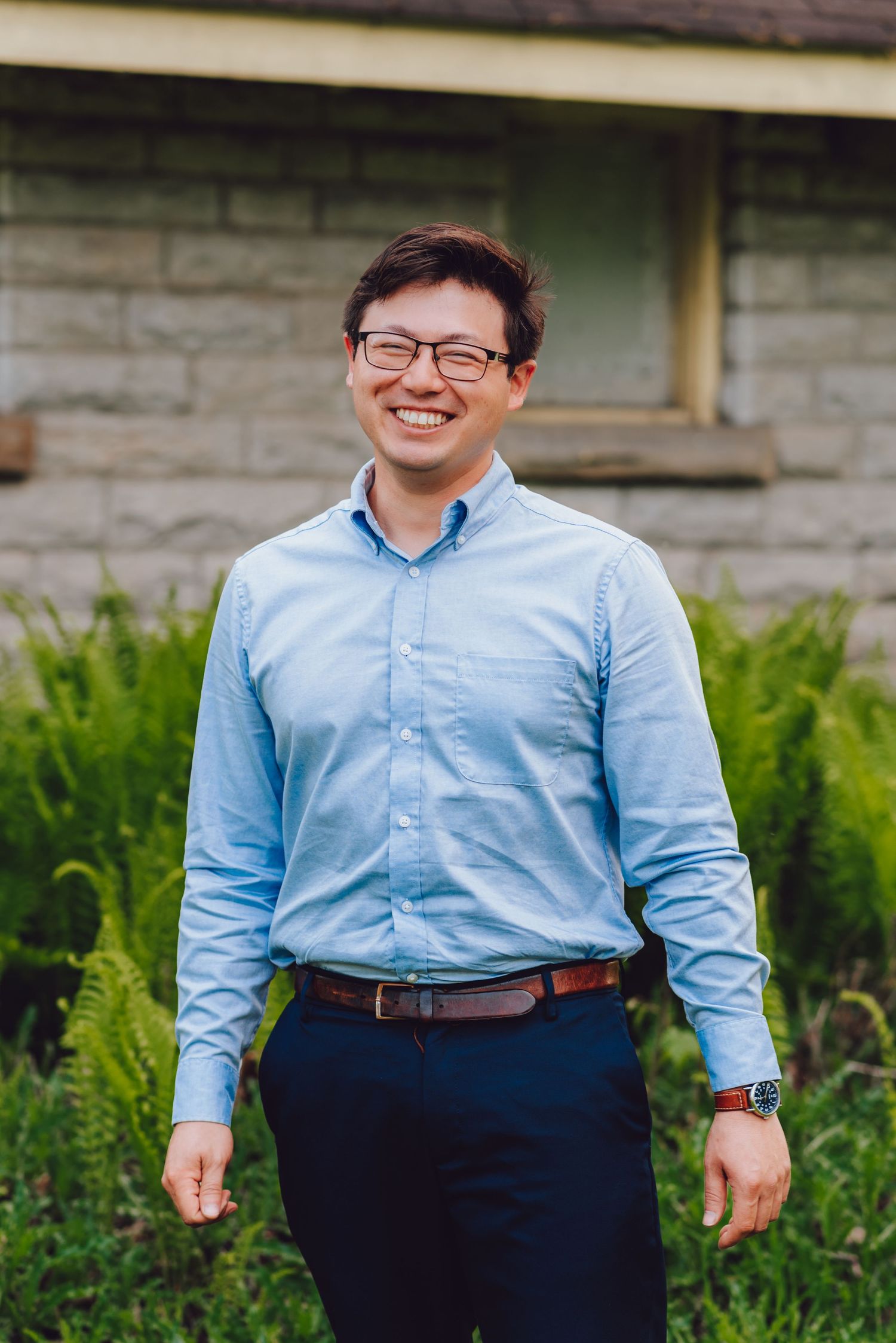 Person in light blue button-down shirt and navy pants standing in front of stone building.