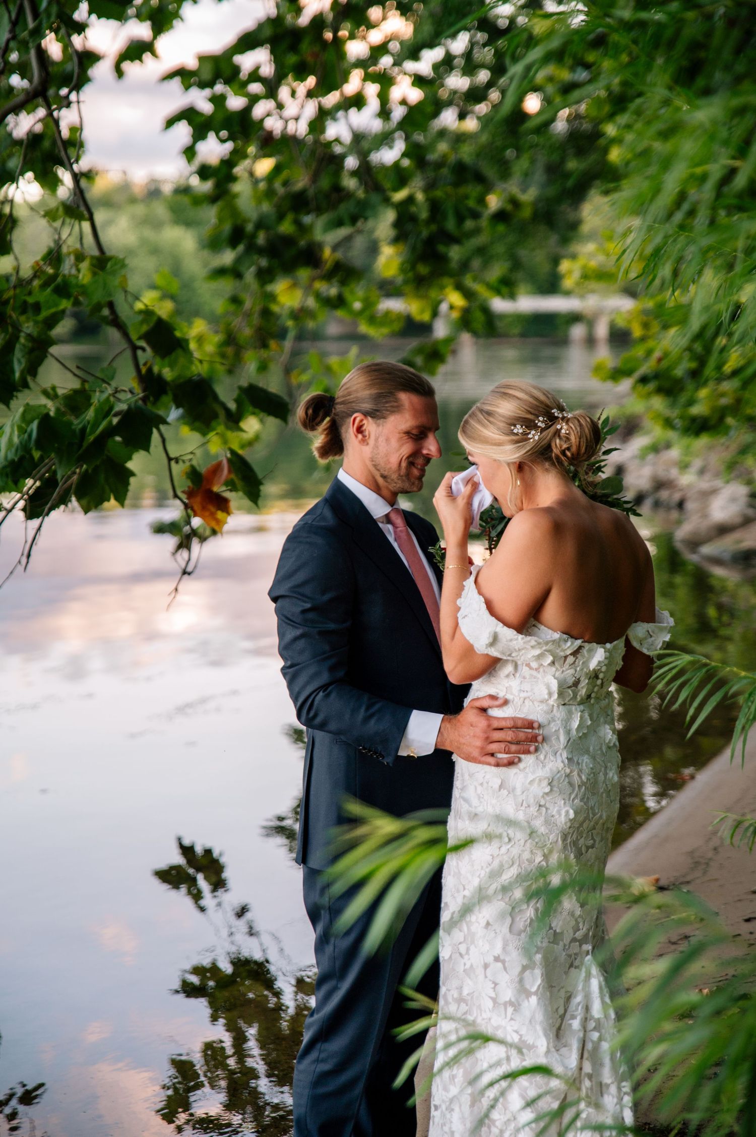 A romantic moment captured between newlyweds by a waterfront with lush foliage.