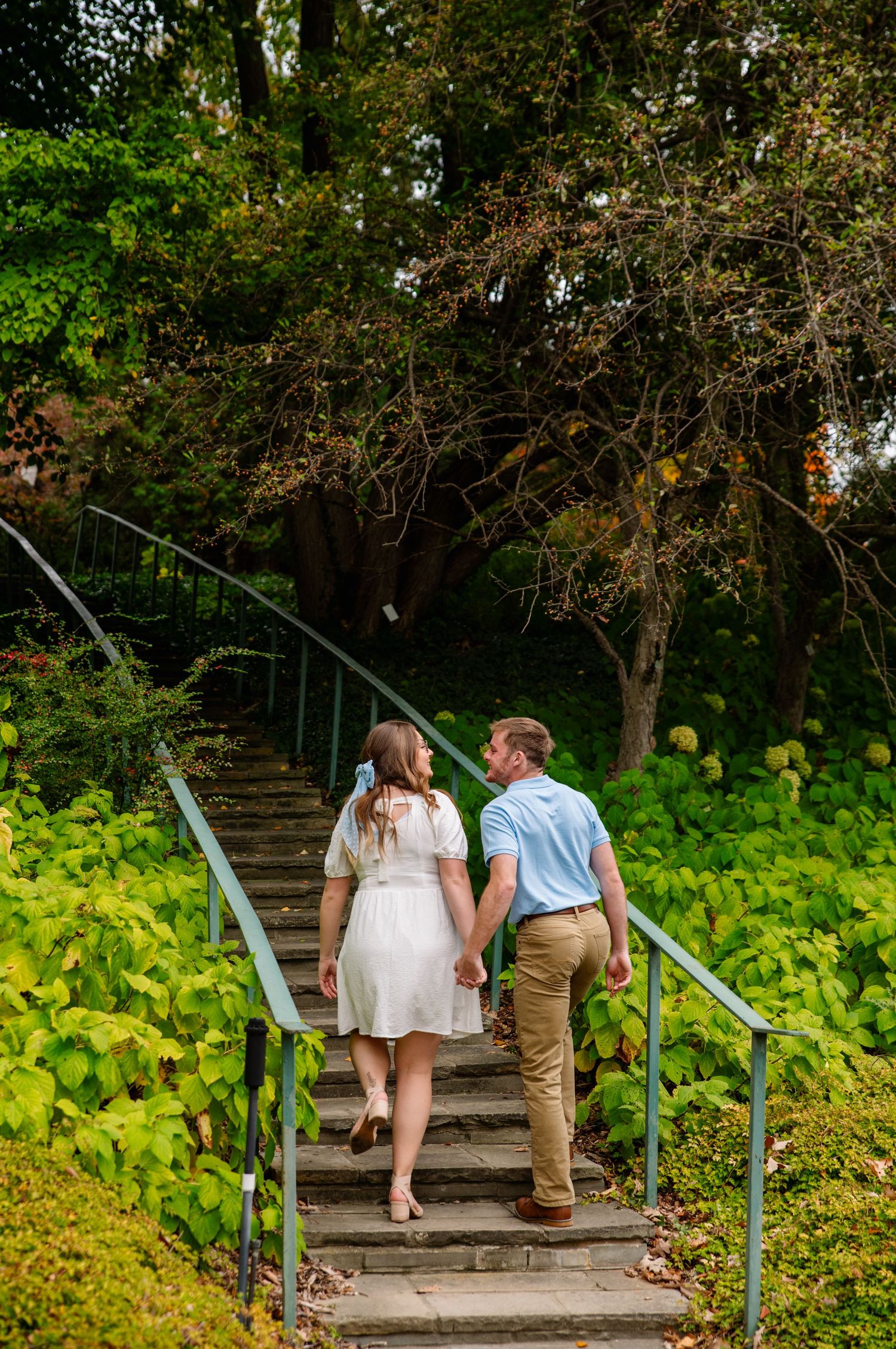 Couple walks up garden steps holding hands surrounded by lush greenery.
