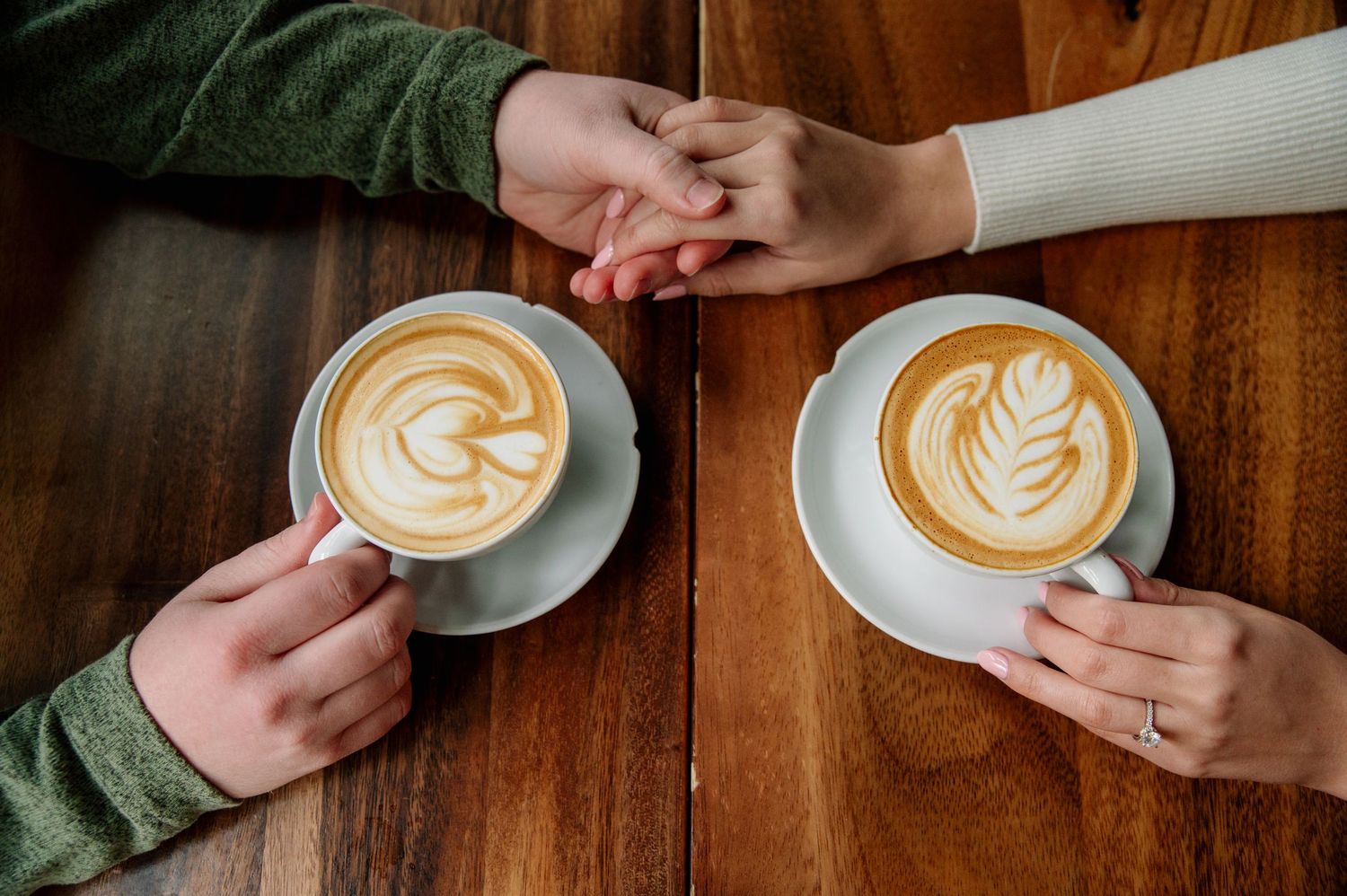 Two hands reach for coffee cups with intricate latte art designs on a wooden table.