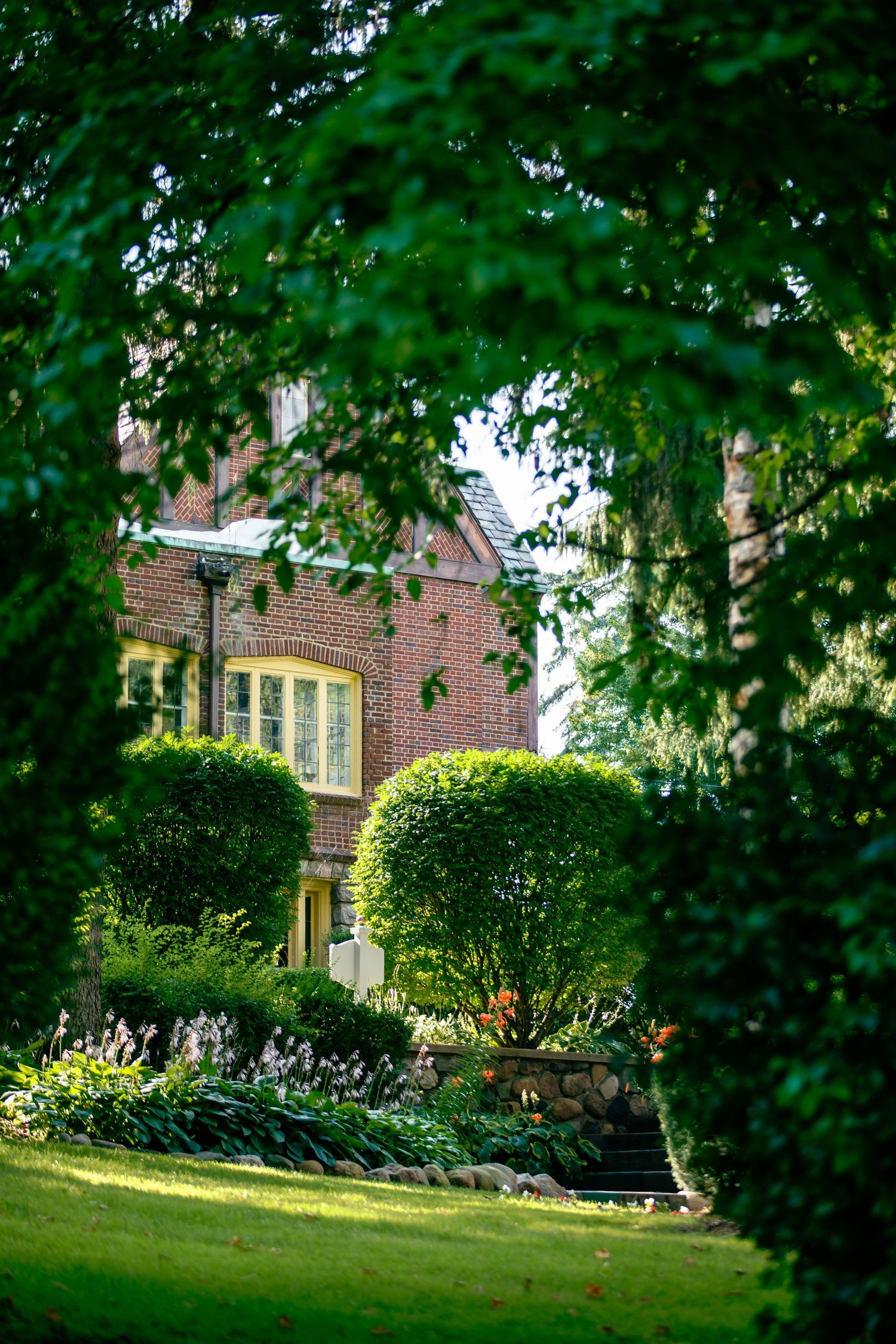 Beautiful landscaped garden with trimmed shrubs and trees framing a traditional brick house on a sunny summer day.