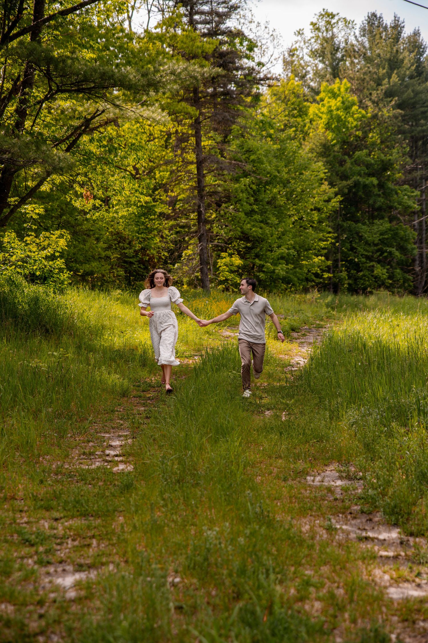 Two people holding hands while walking along a grassy path through a sun-drenched forest.