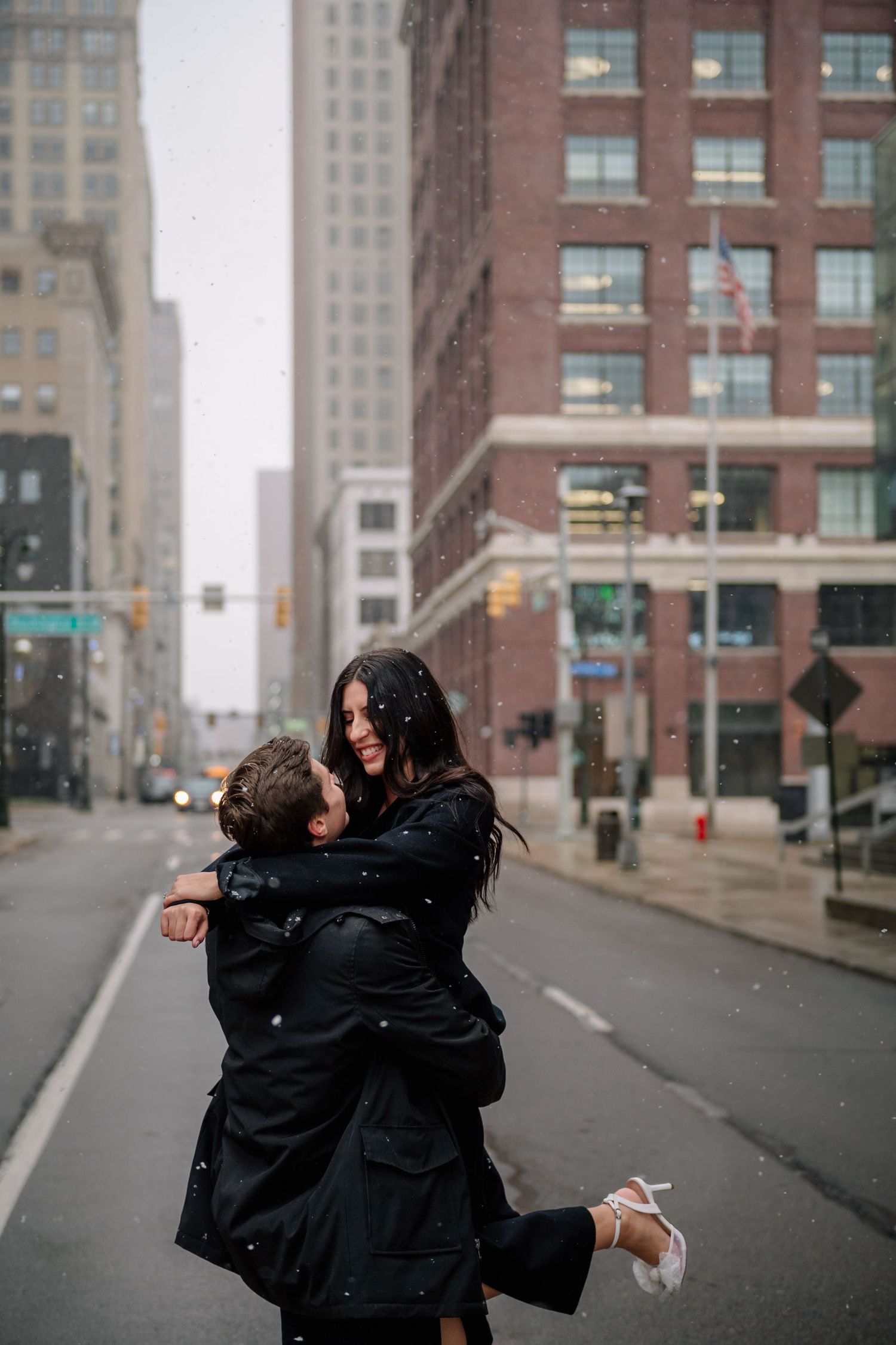 A joyful couple spins in an embrace on a snowy city street surrounded by tall buildings.
