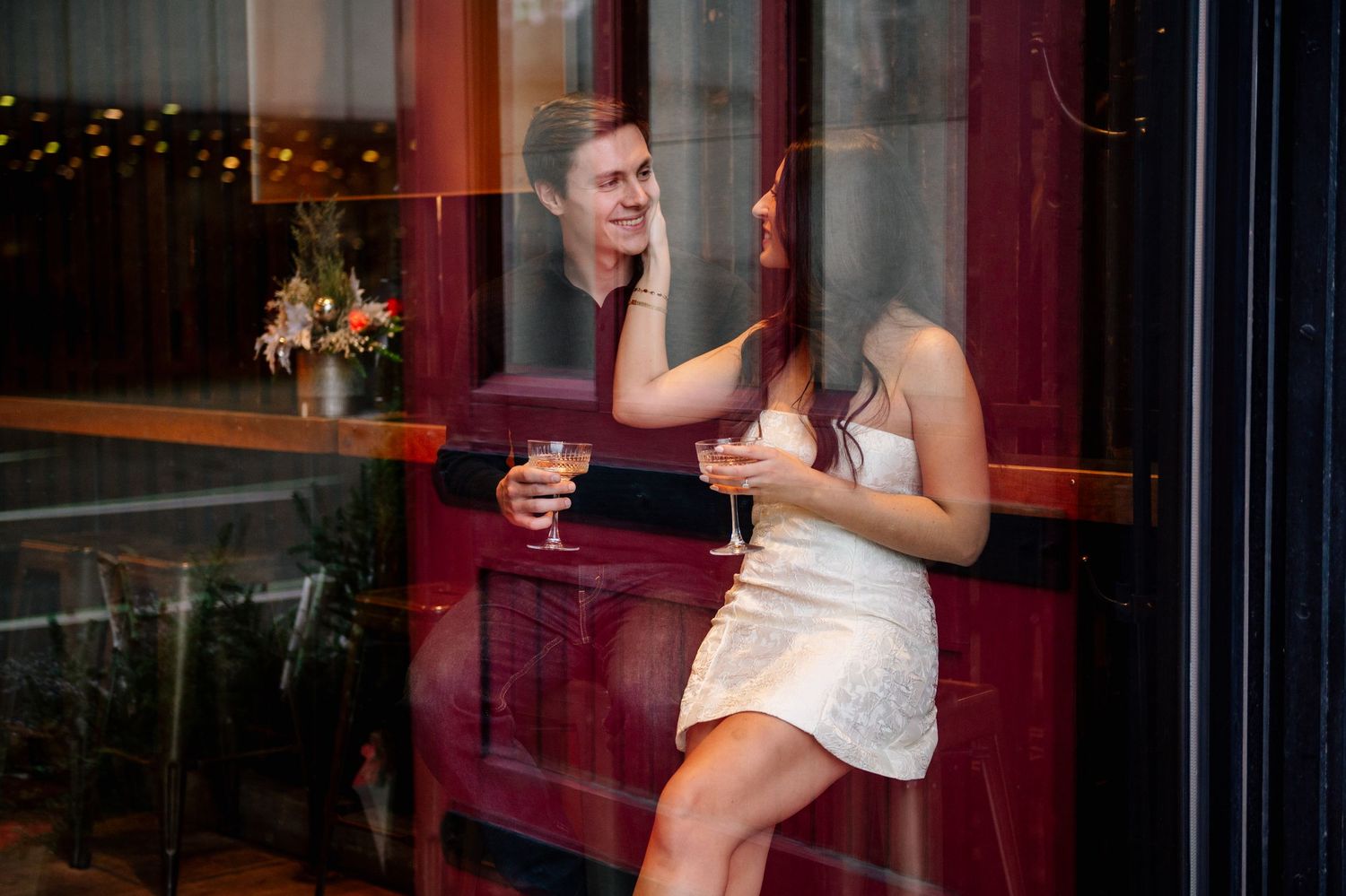 A couple shares a laugh while enjoying drinks by a window in an evening setting.