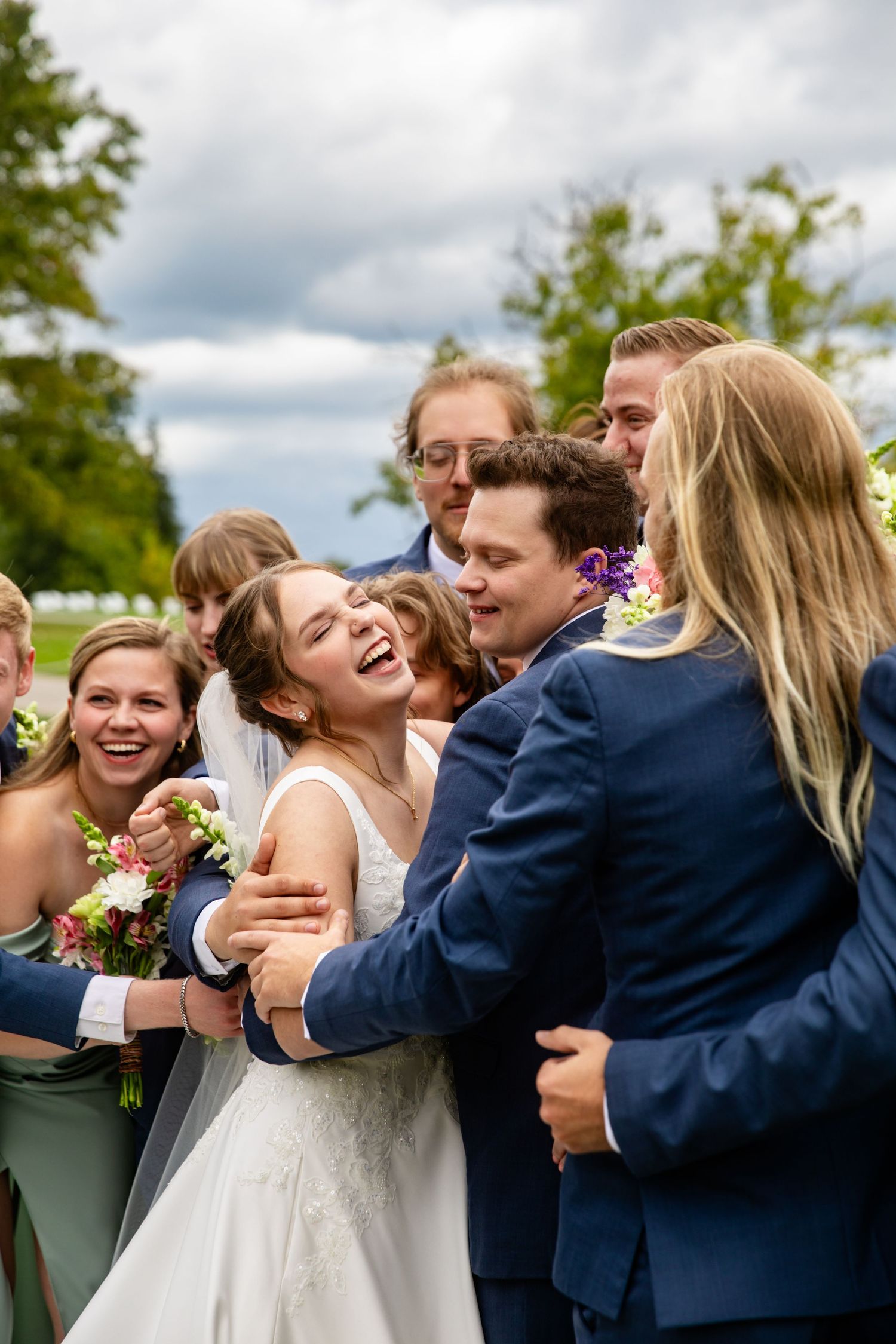 A joyful wedding party laughs together outdoors on a cloudy day with greenery in the background.