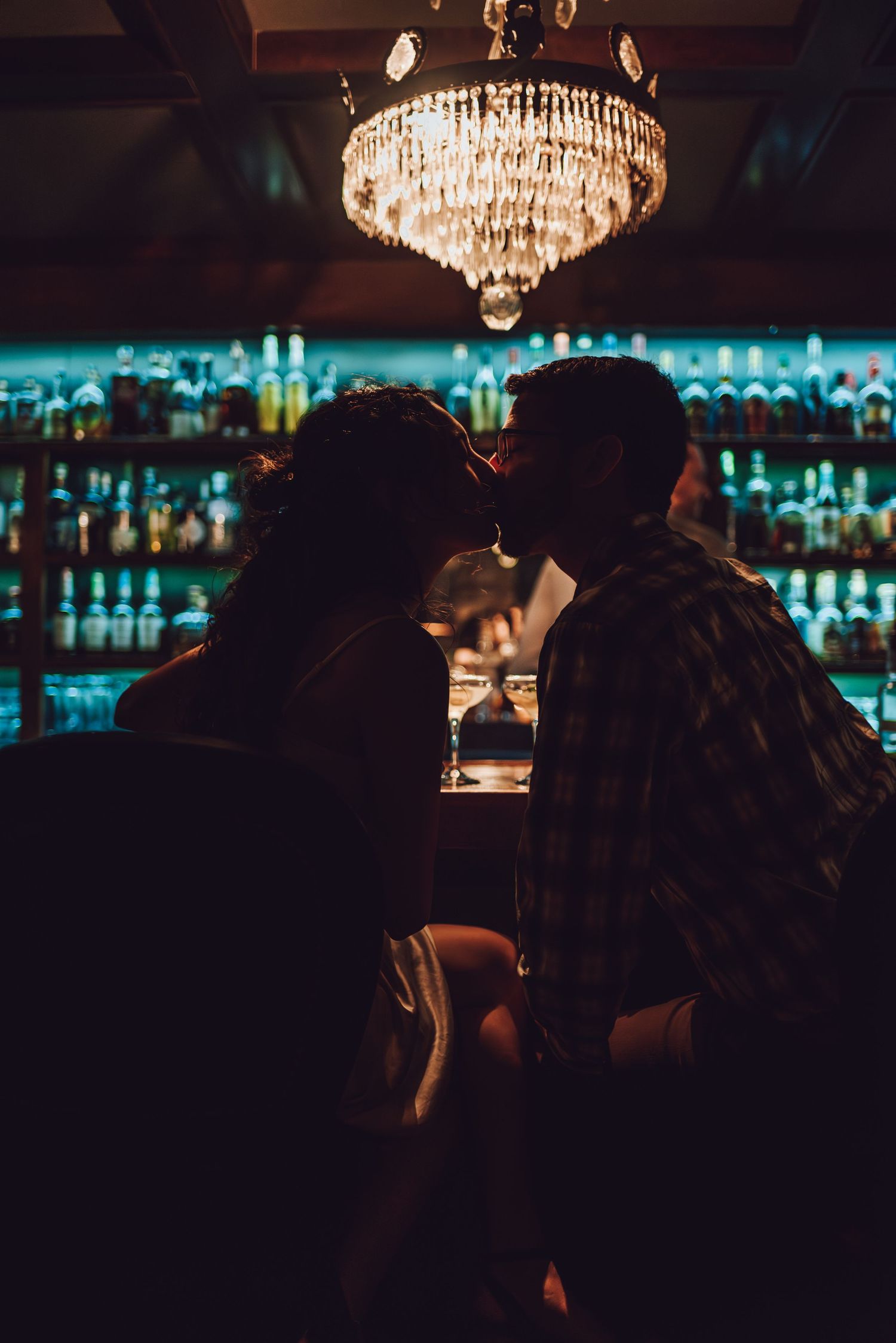 Silhouetted couple sharing intimate moment under crystal chandelier at dimly lit bar.