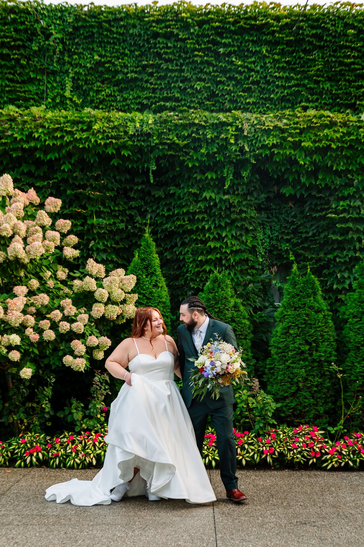 Wedding couple shares romantic moments in front of lush garden wall with peach hydrangeas and red flowers.