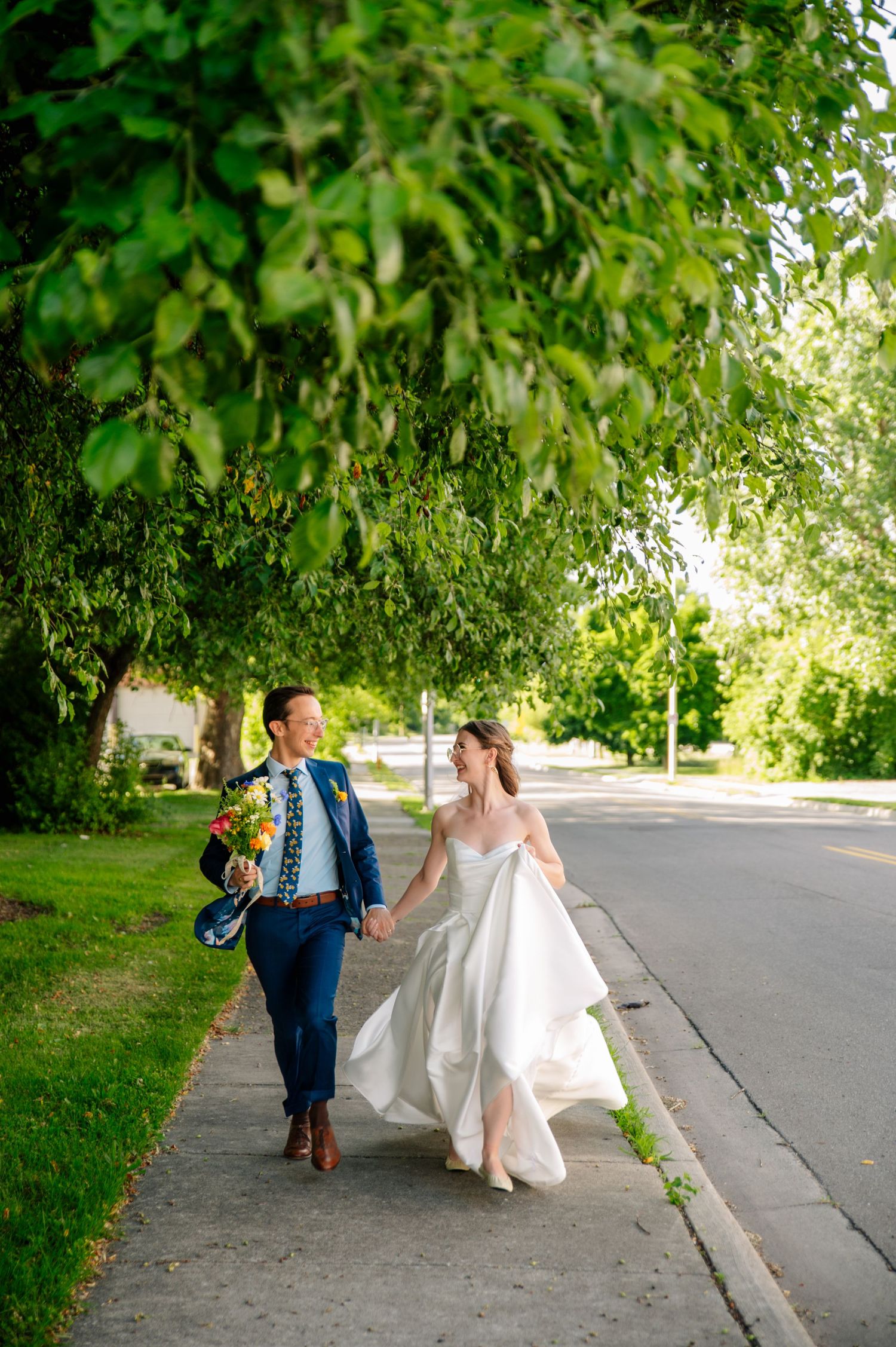A couple walks joyfully along a tree-lined path on their wedding day in summer.