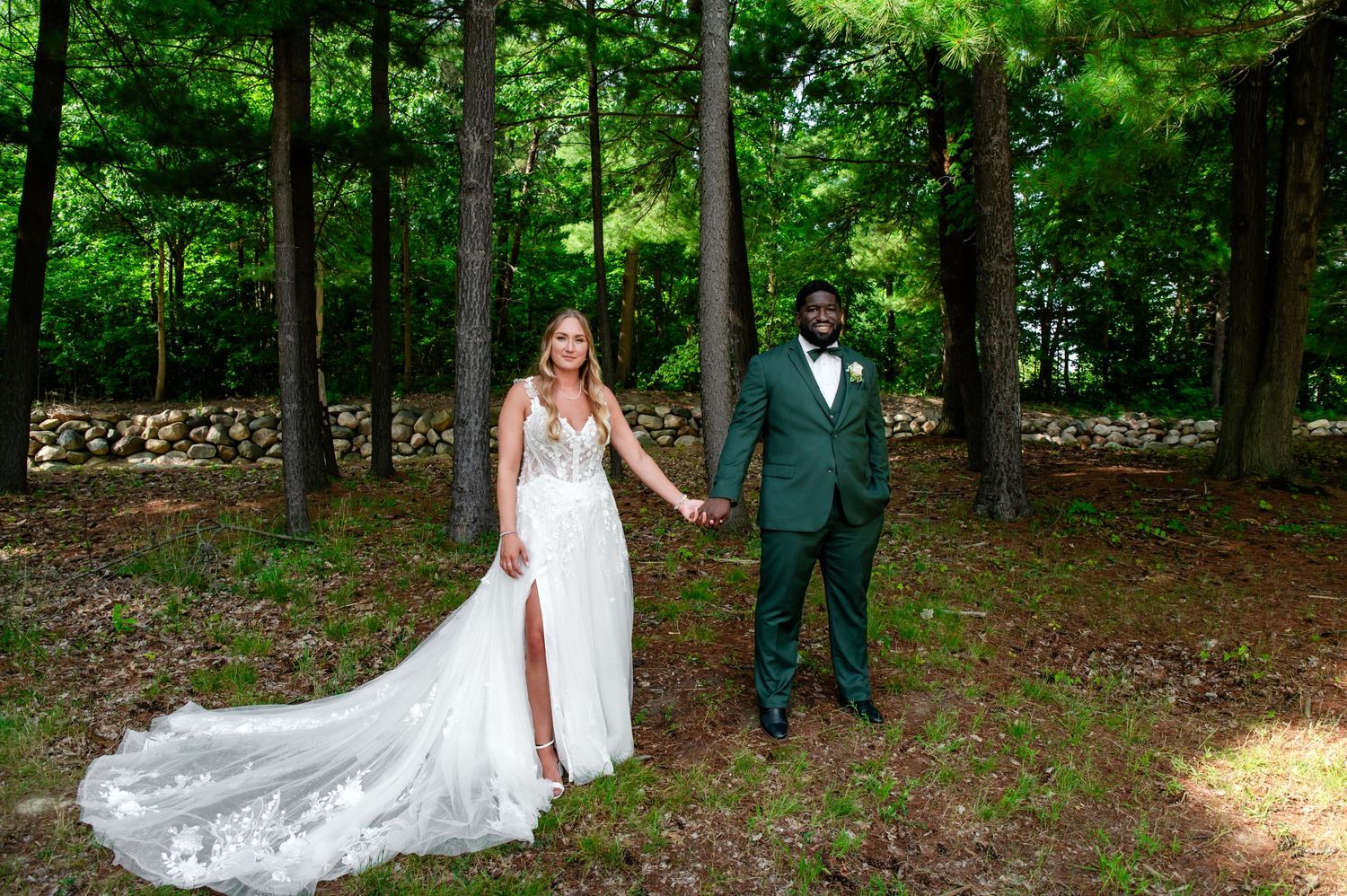 A bride in a flowing white wedding dress walks through a sunlit wooded area during an outdoor photo session.